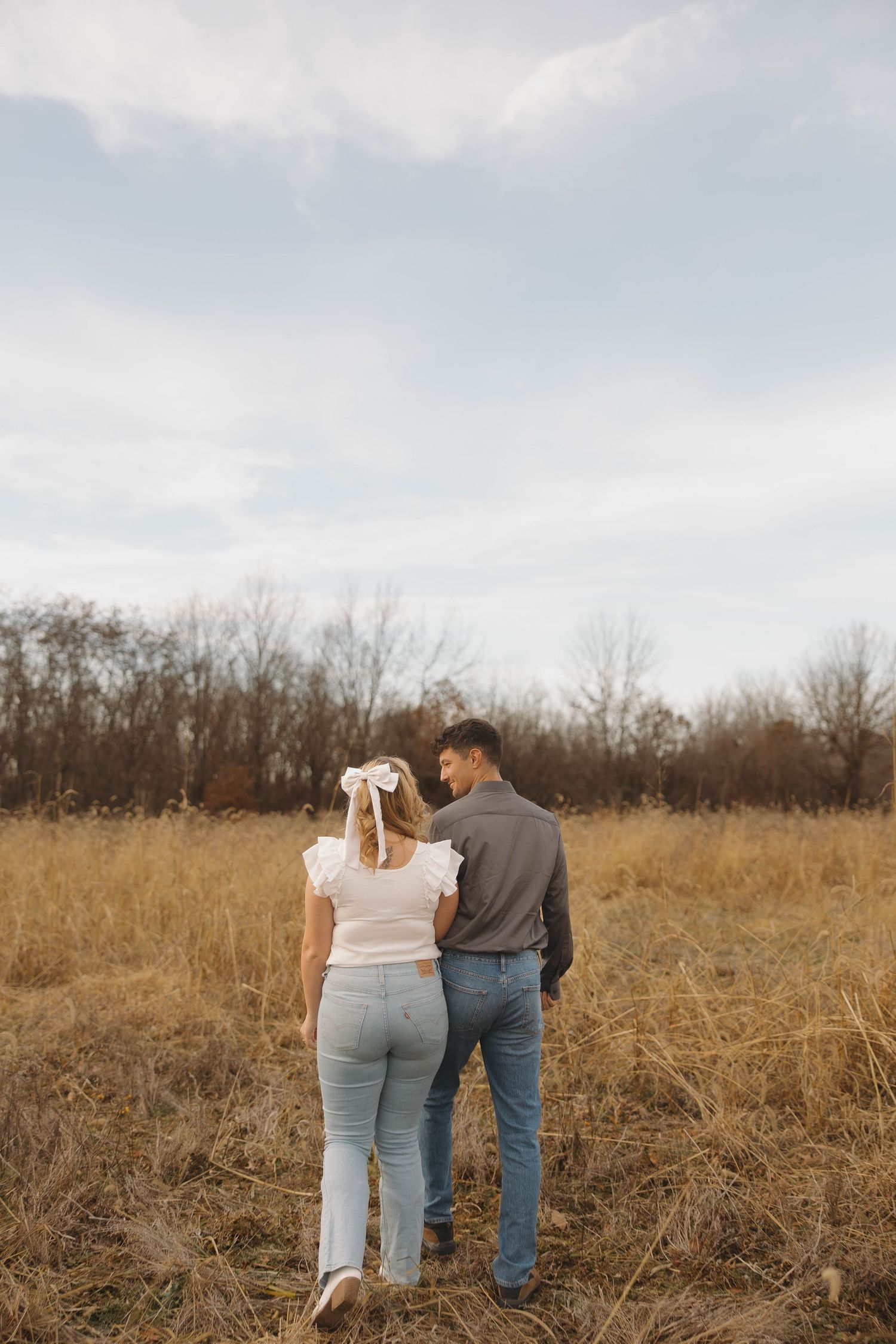 A couple stands together in a golden field on an autumn day with bare trees in the background.