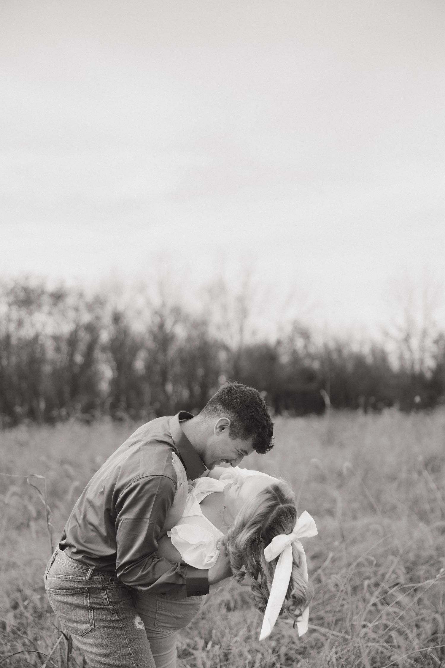 A person in vintage clothing bends down to pet a small white dog in a grassy field.