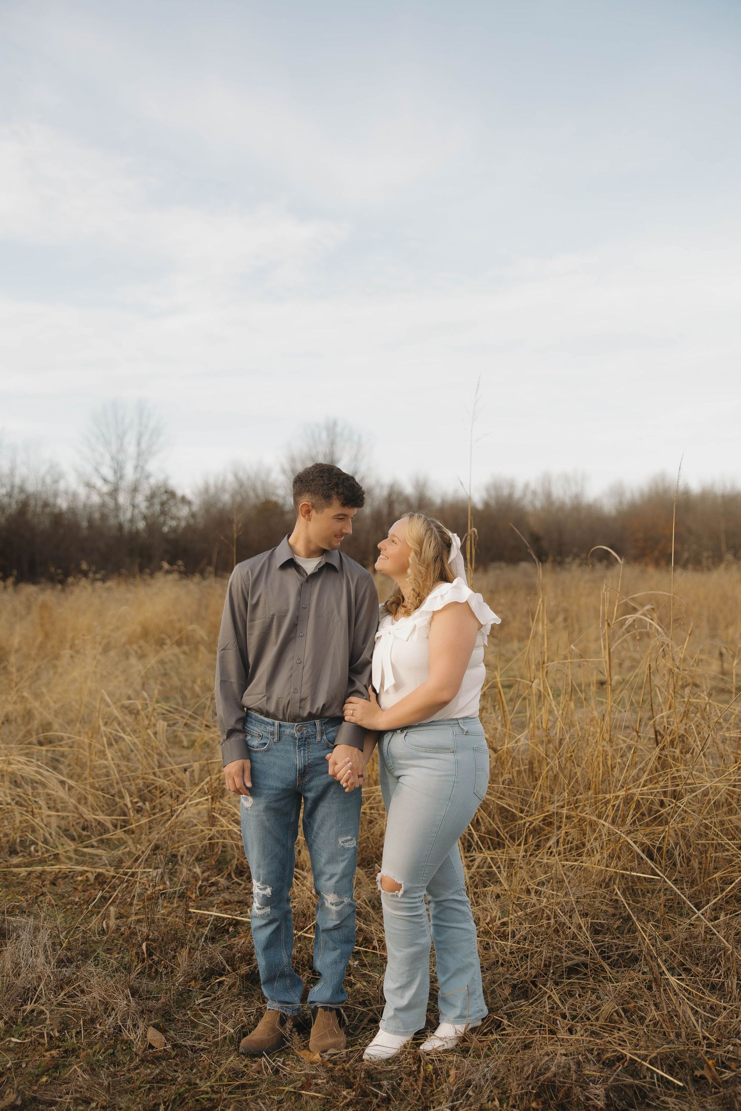 A couple stands together in a golden autumn field wearing casual denim and light colored clothing.