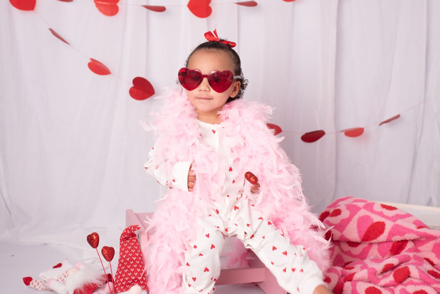 Small child in pink tulle outfit and heart sunglasses poses with Valentine's Day decorations and falling rose petals.