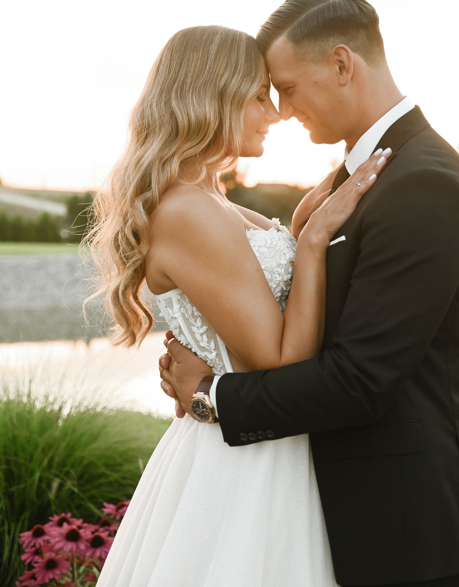 Romantic newlywed couple embracing in golden sunlight by a garden with pink flowers at sunset.
