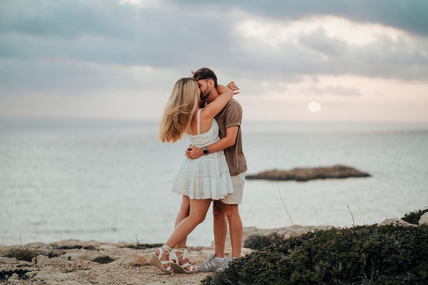 A couple embraces on a rocky beach during sunset with dramatic cloudy skies over the ocean.