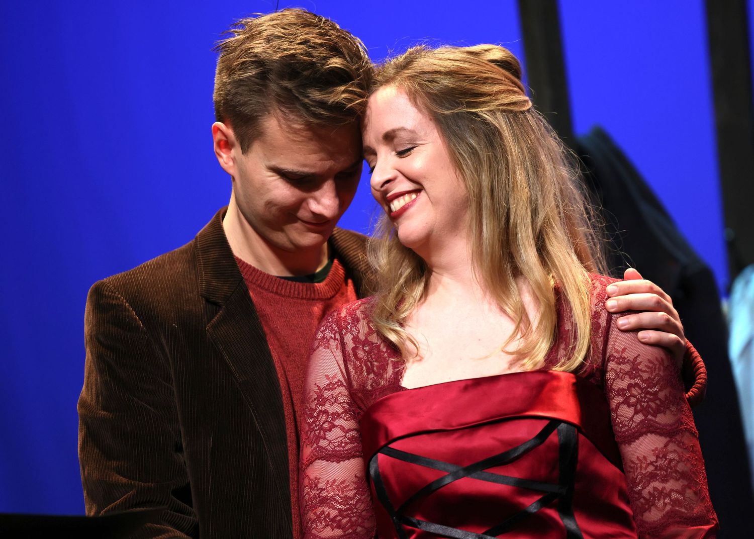 A couple shares an intimate moment while performing together on stage against a blue backdrop.