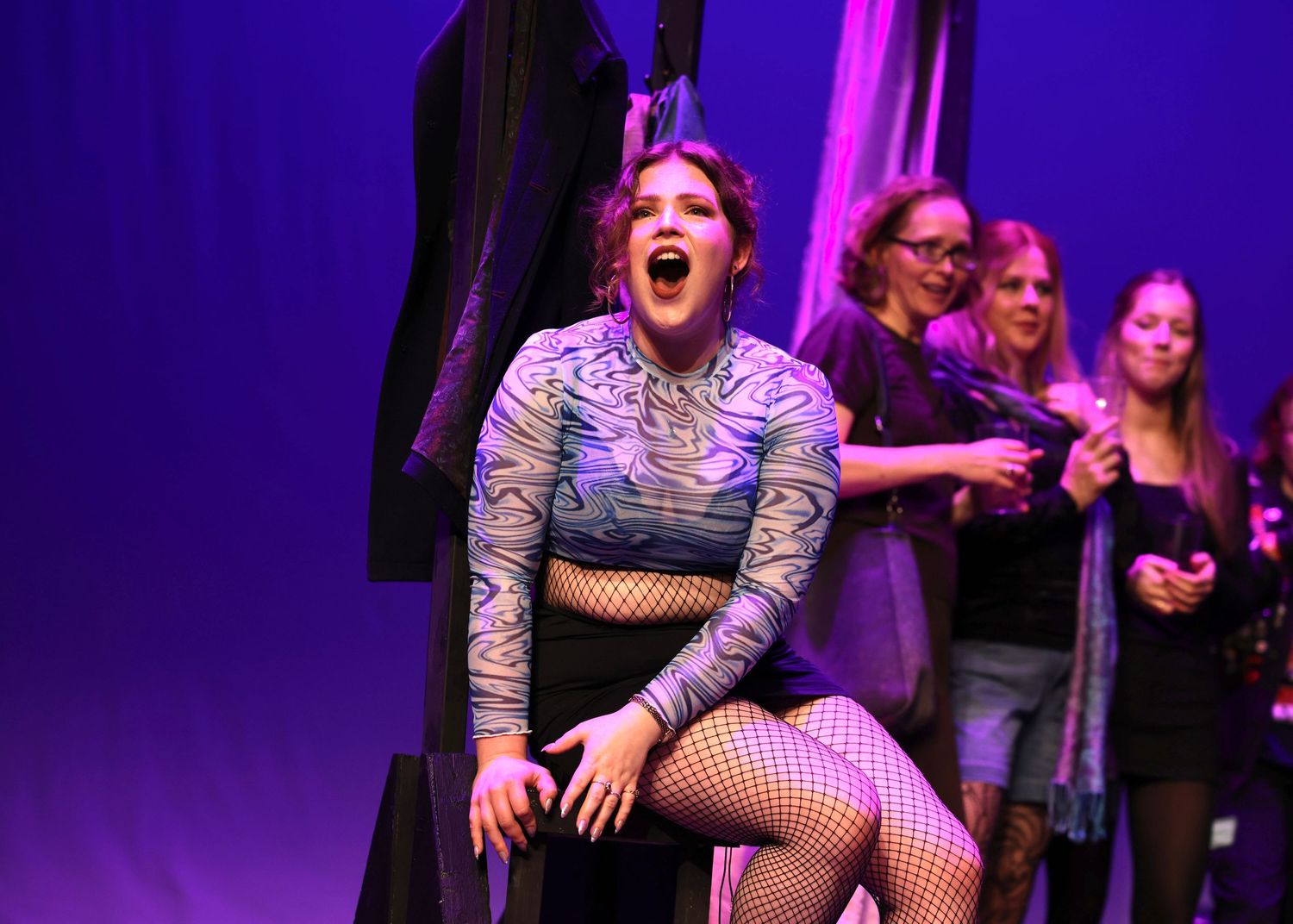 Performers in theatrical costumes dance on a purple-lit stage during an energetic musical theater production.