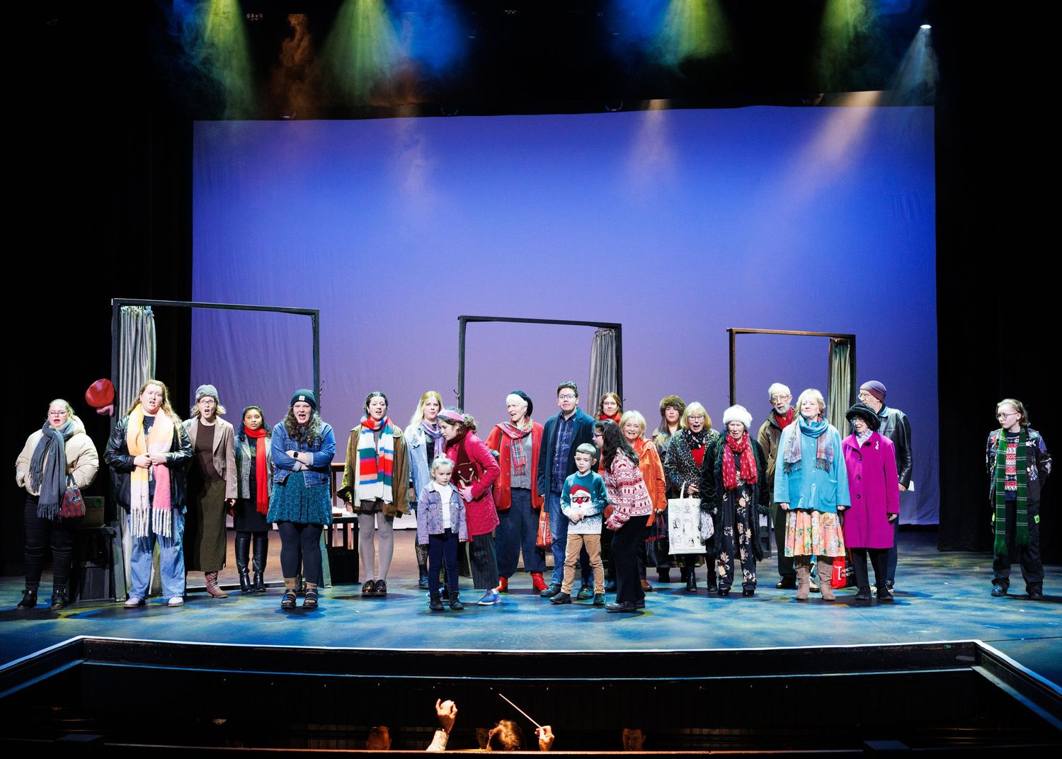 A theatrical stage performance with a large cast in winter attire against a blue-lit backdrop.