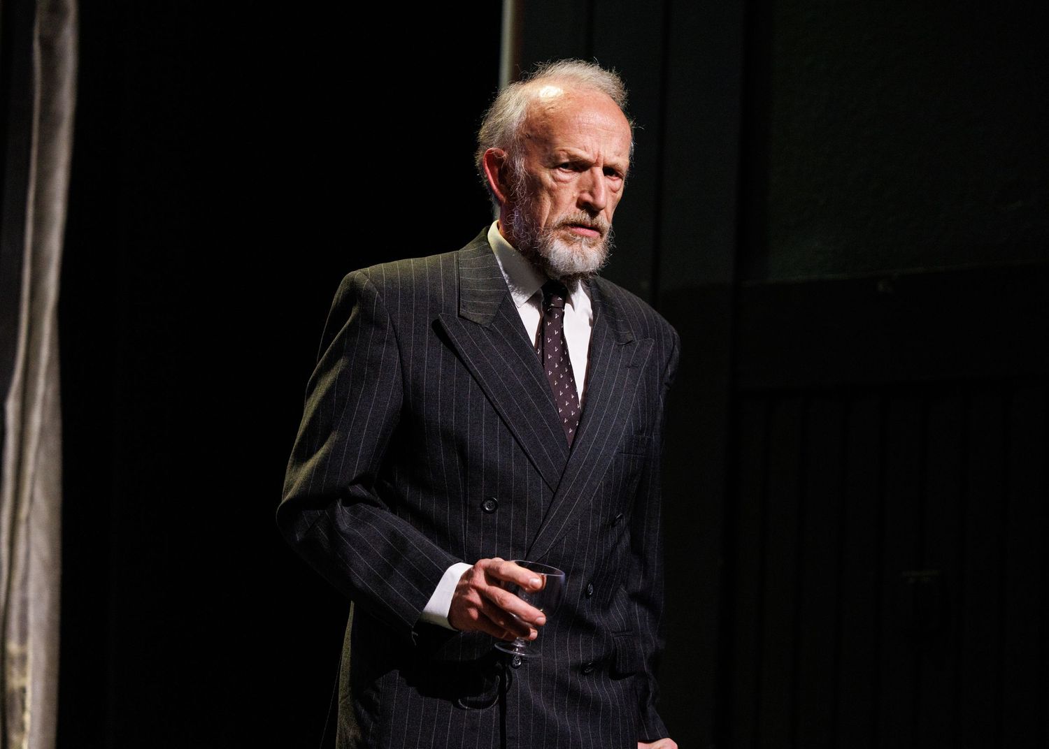 A performer in a pinstriped suit gestures dramatically on a dark theater stage during a performance.