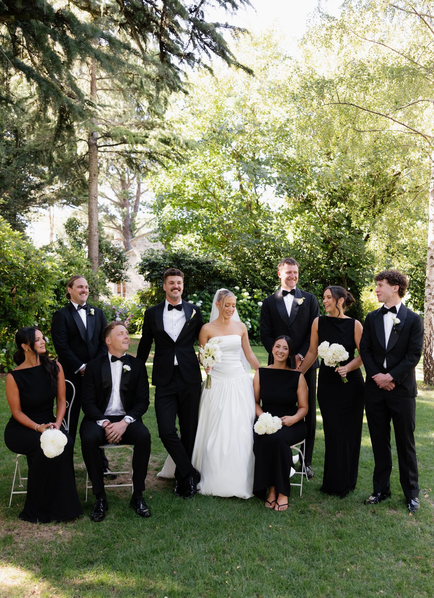 A wedding party in black formal attire and white gowns poses together on a grassy lawn with trees in the background.