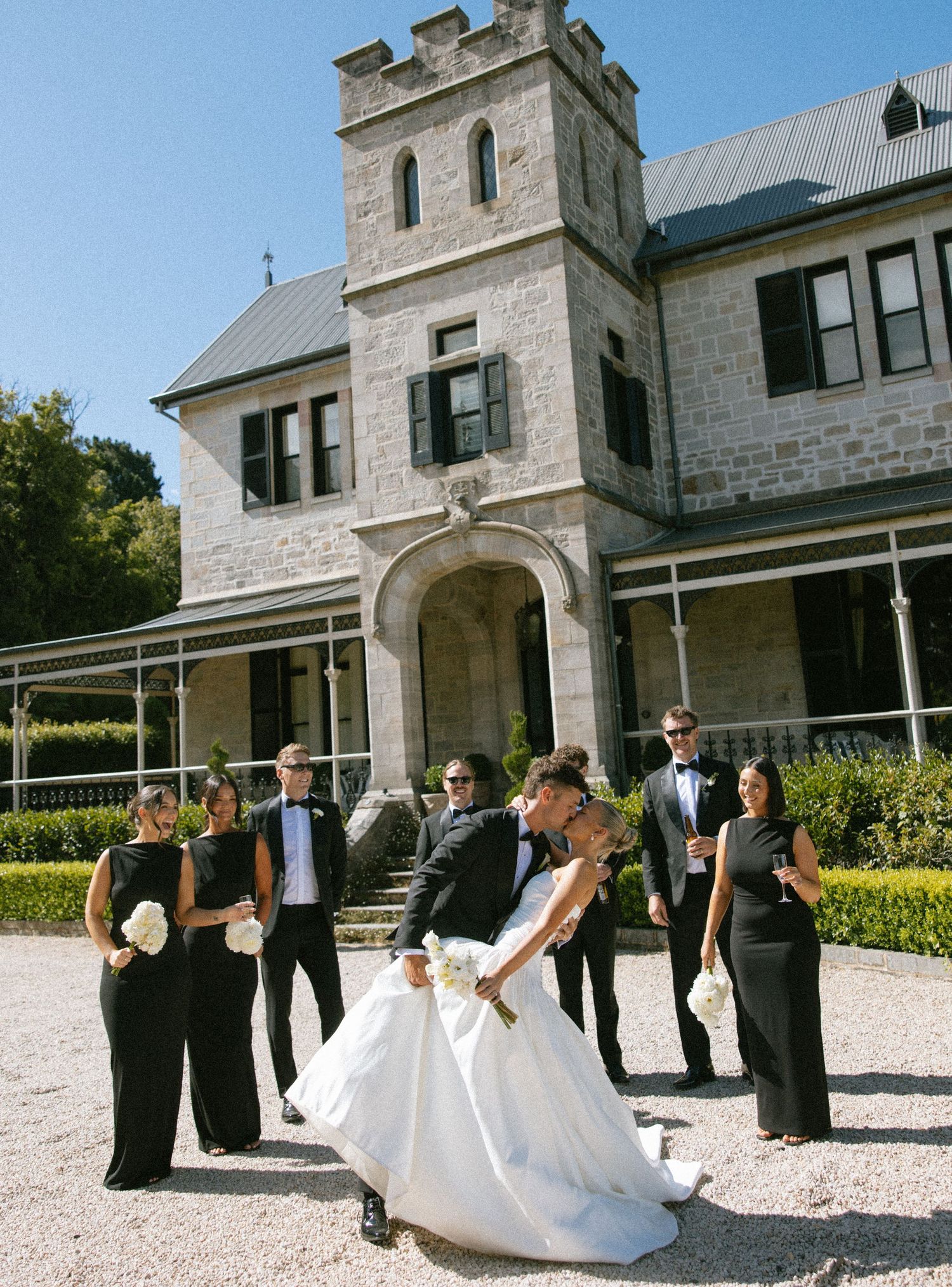 Wedding party members in black attire pose playfully with a bride in a white gown outside a stone building.