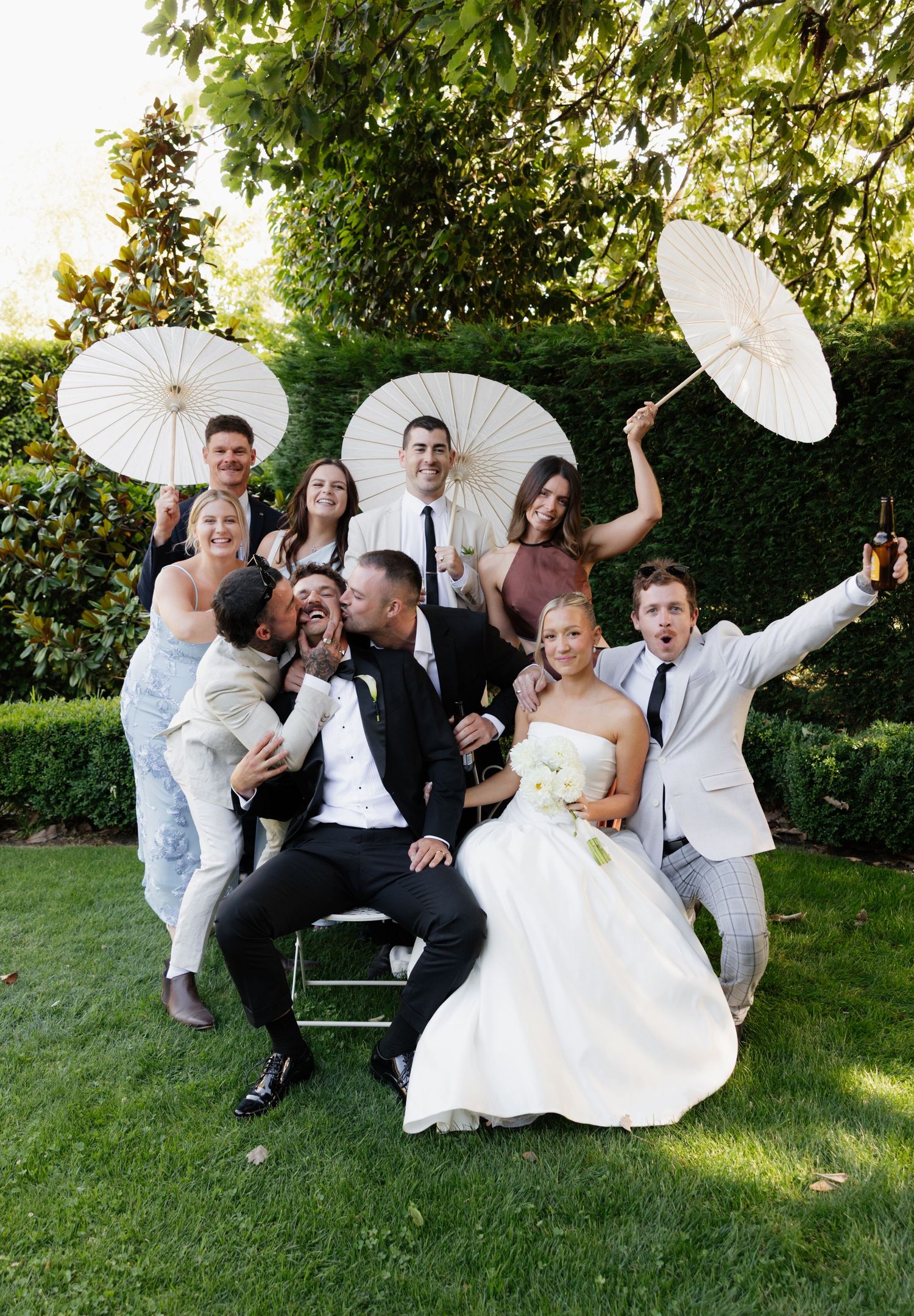 A wedding party poses playfully with white parasols in a garden setting during a sunny outdoor celebration.