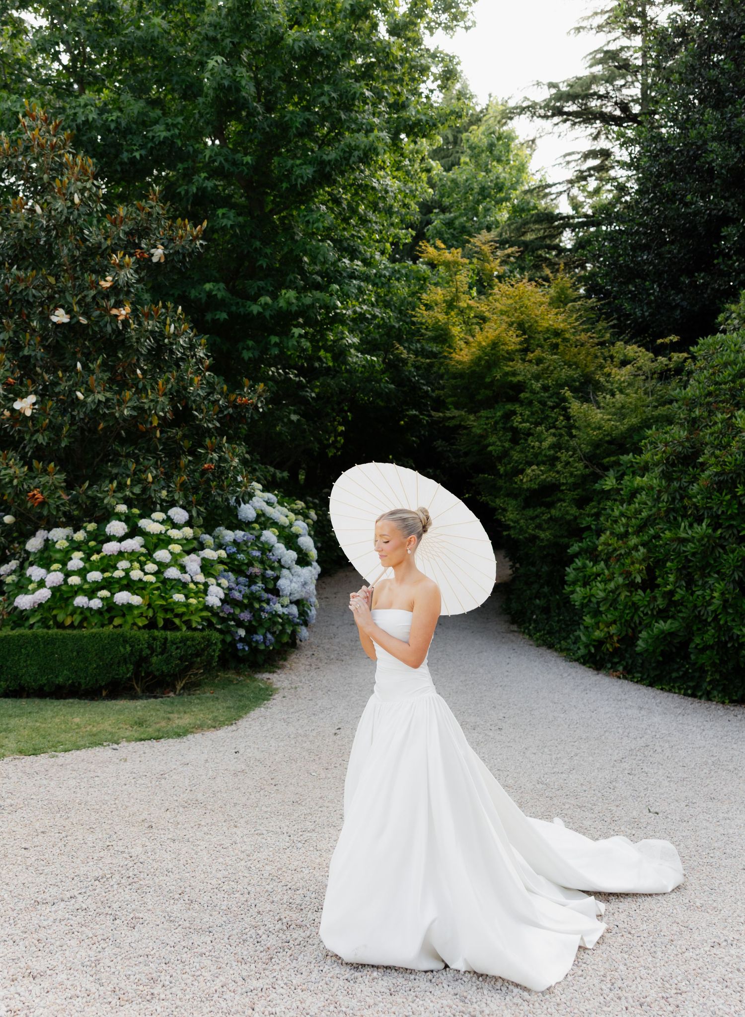 A bride in a flowing white gown twirls gracefully on a garden path surrounded by lush greenery and white hydrangeas.