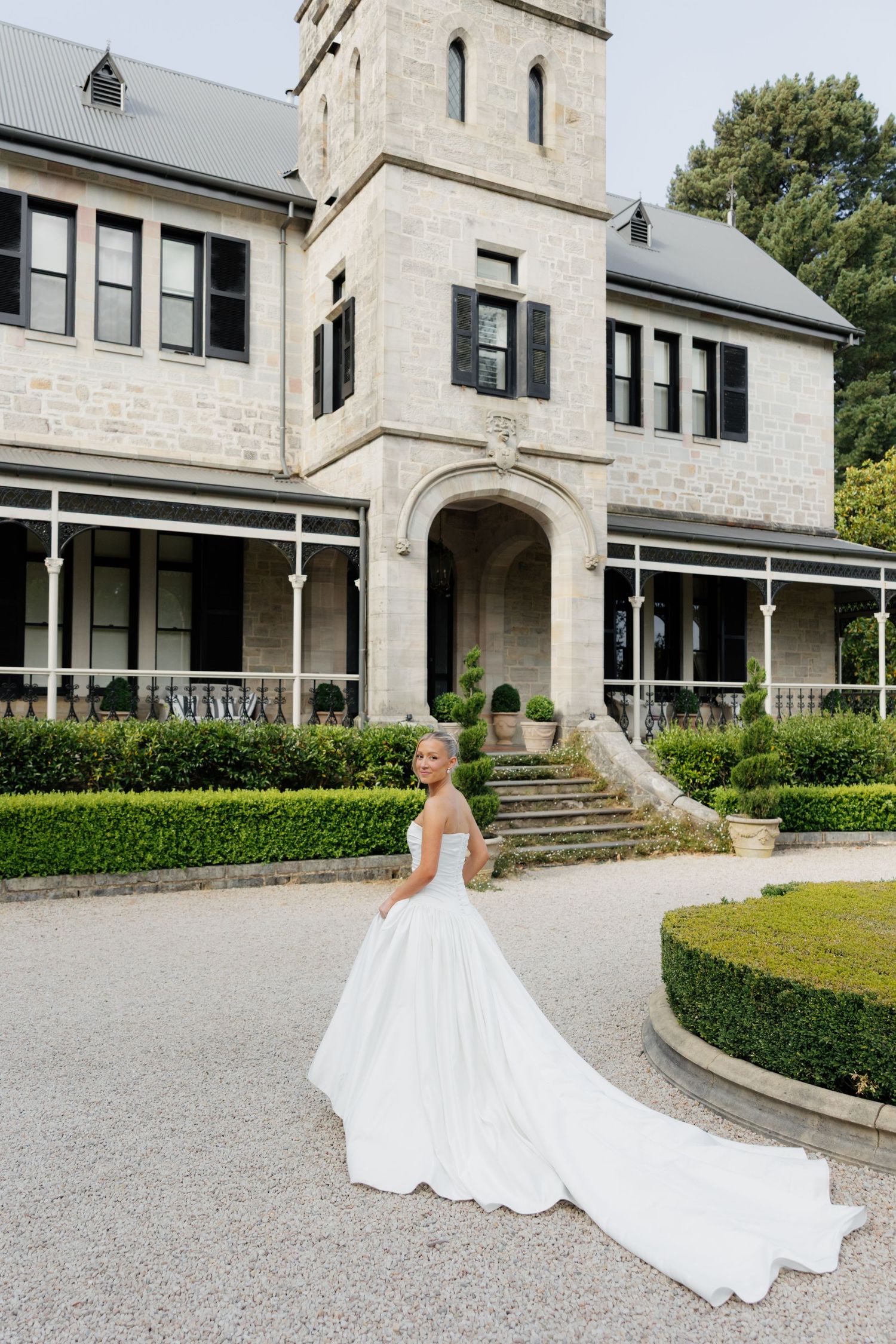 A stunning white wedding dress with a long train photographed against the backdrop of a stone mansion and manicured gardens.