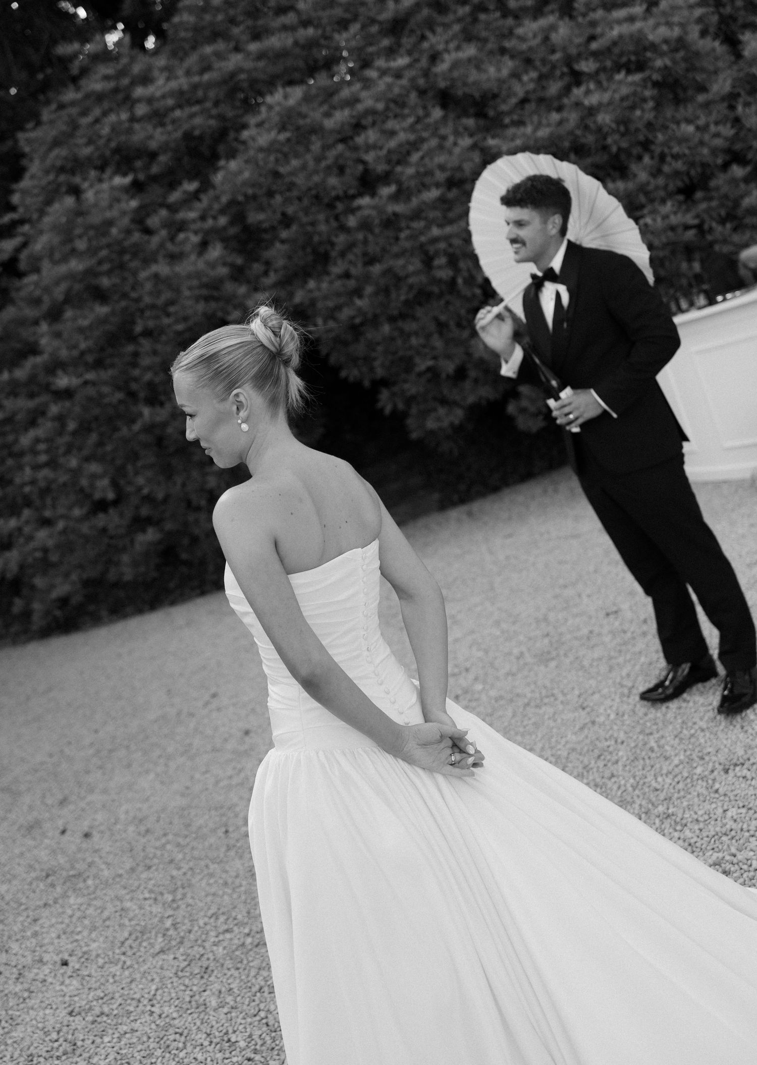 Bride in strapless gown adjusts her dress train on gravel path surrounded by lush garden foliage.