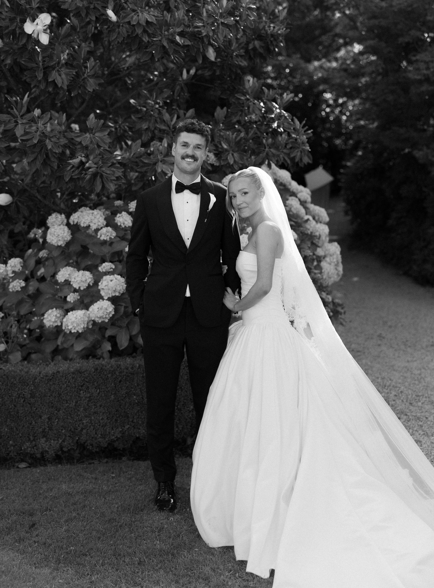 A black and white wedding photo captures a romantic moment between newlyweds in a garden setting with hydrangeas.