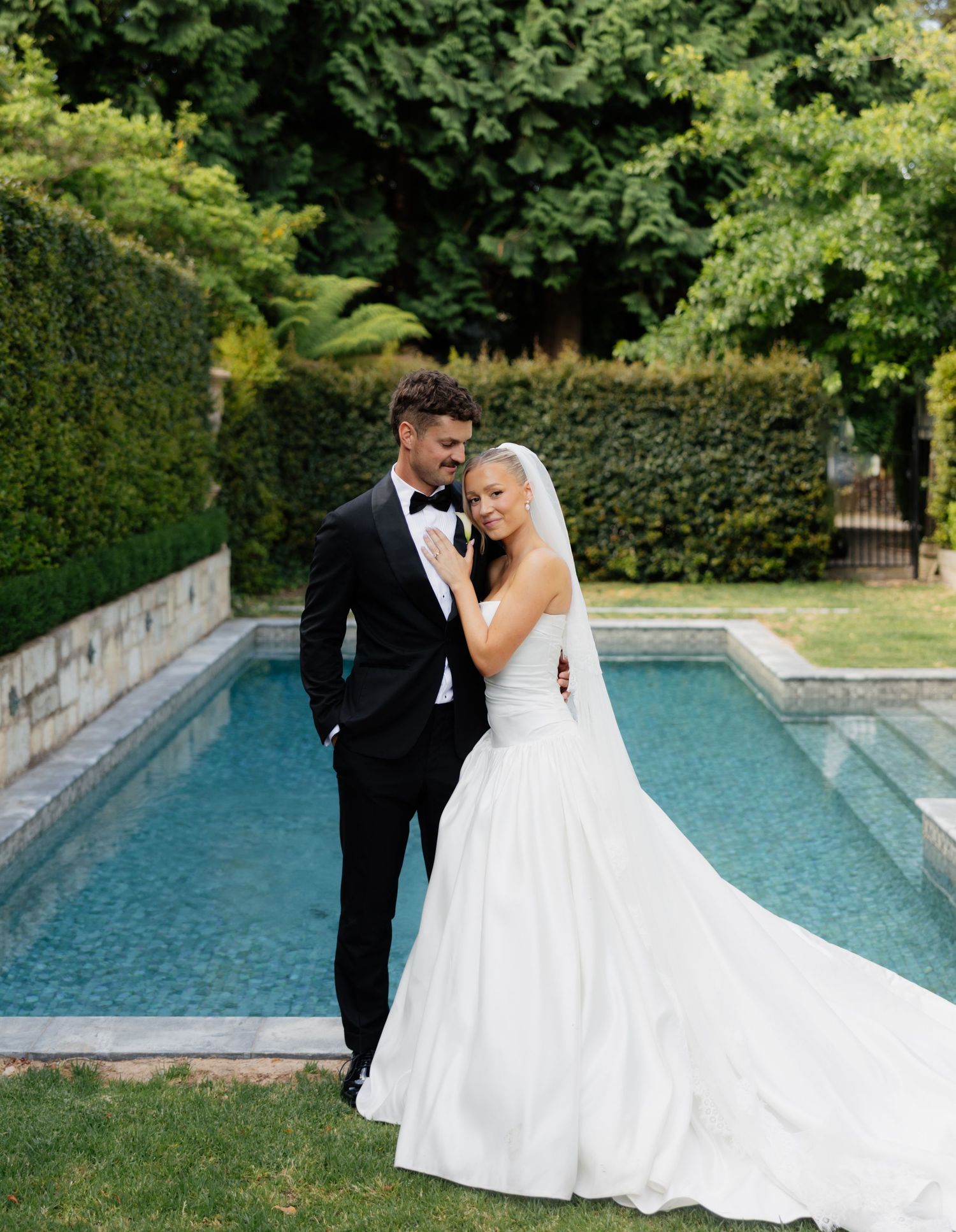 A couple shares an intimate moment by a tranquil swimming pool surrounded by lush greenery on their wedding day.