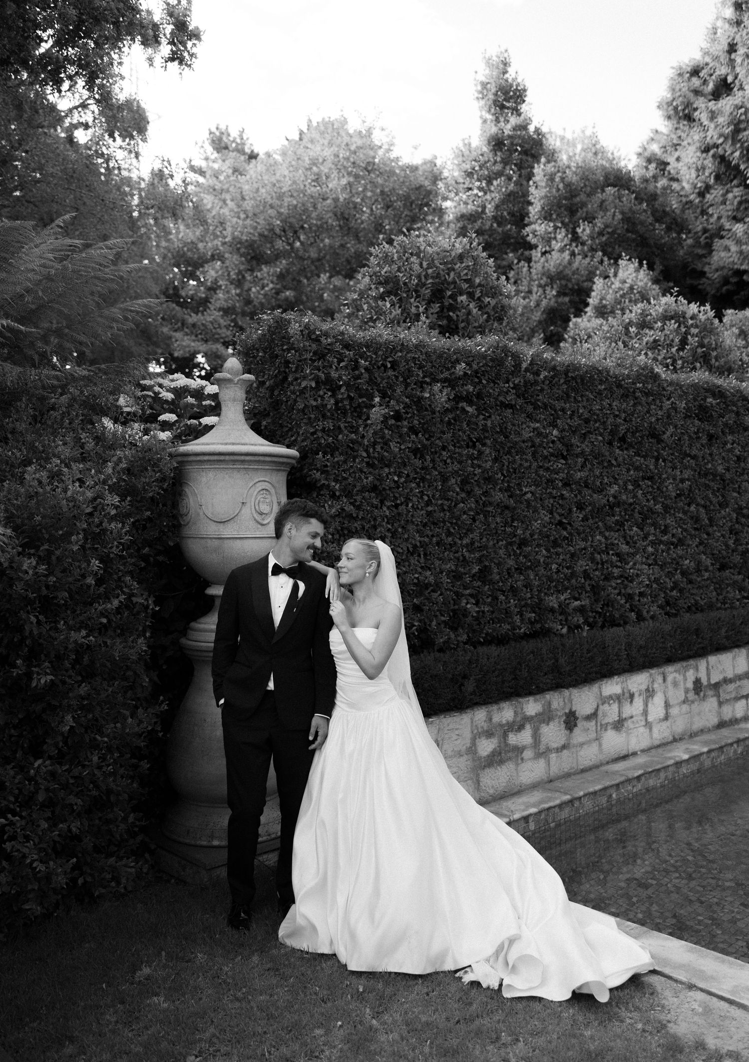 A romantic black and white wedding photo series showing a couple walking along a stone wall lined with hedges.