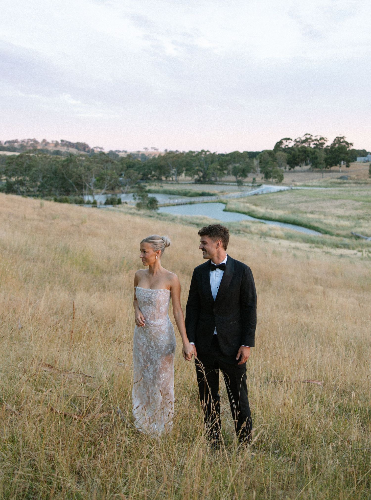 Newlyweds pose together in an open field with rolling hills and golden grass in the background.