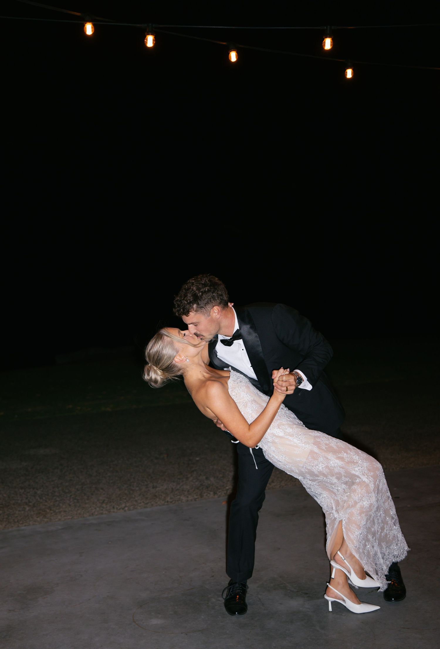Couple dancing romantically under string lights at night during their wedding reception.