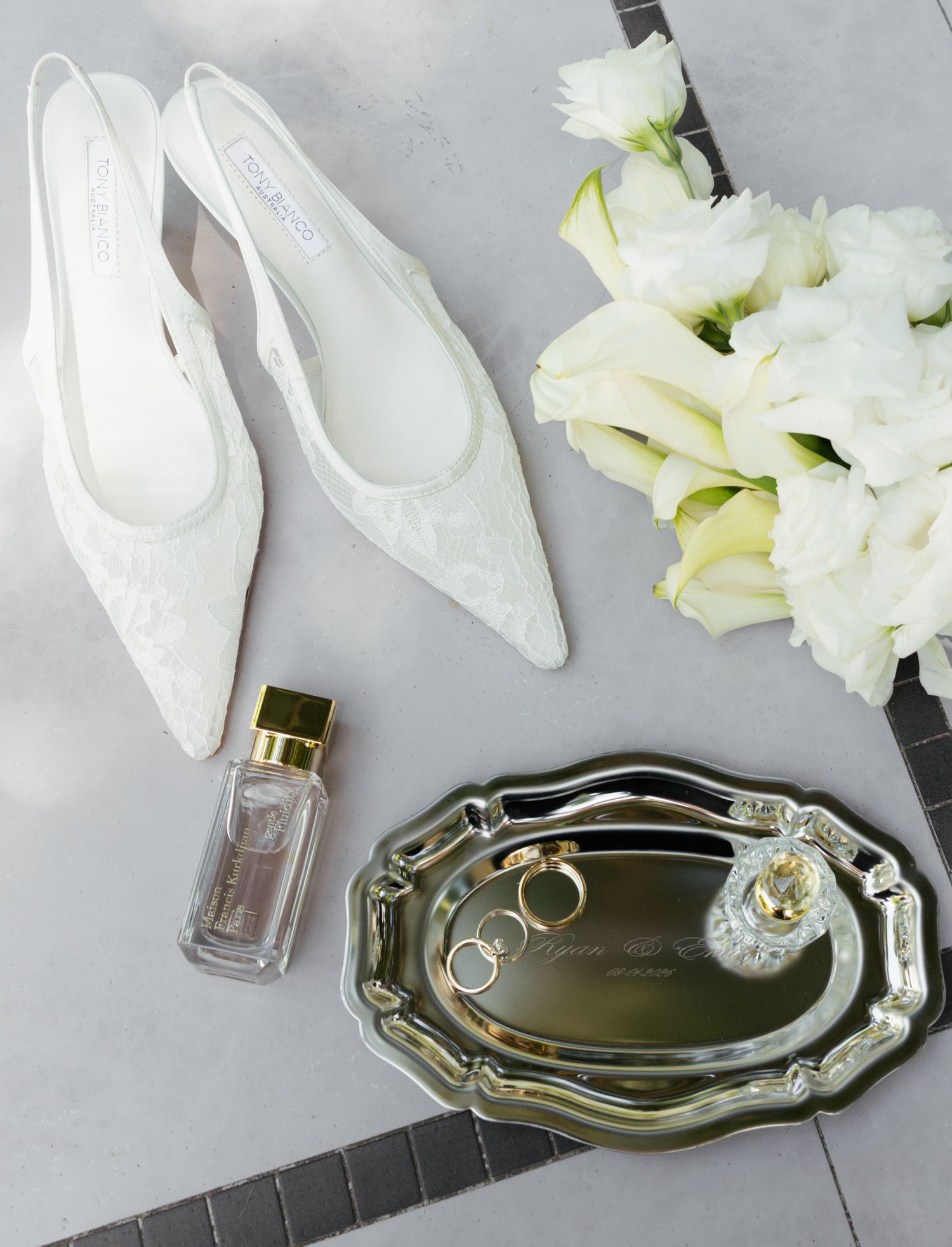 Elegant white bridal shoes and flowers arranged with silver trays and perfume bottles on a gray background.