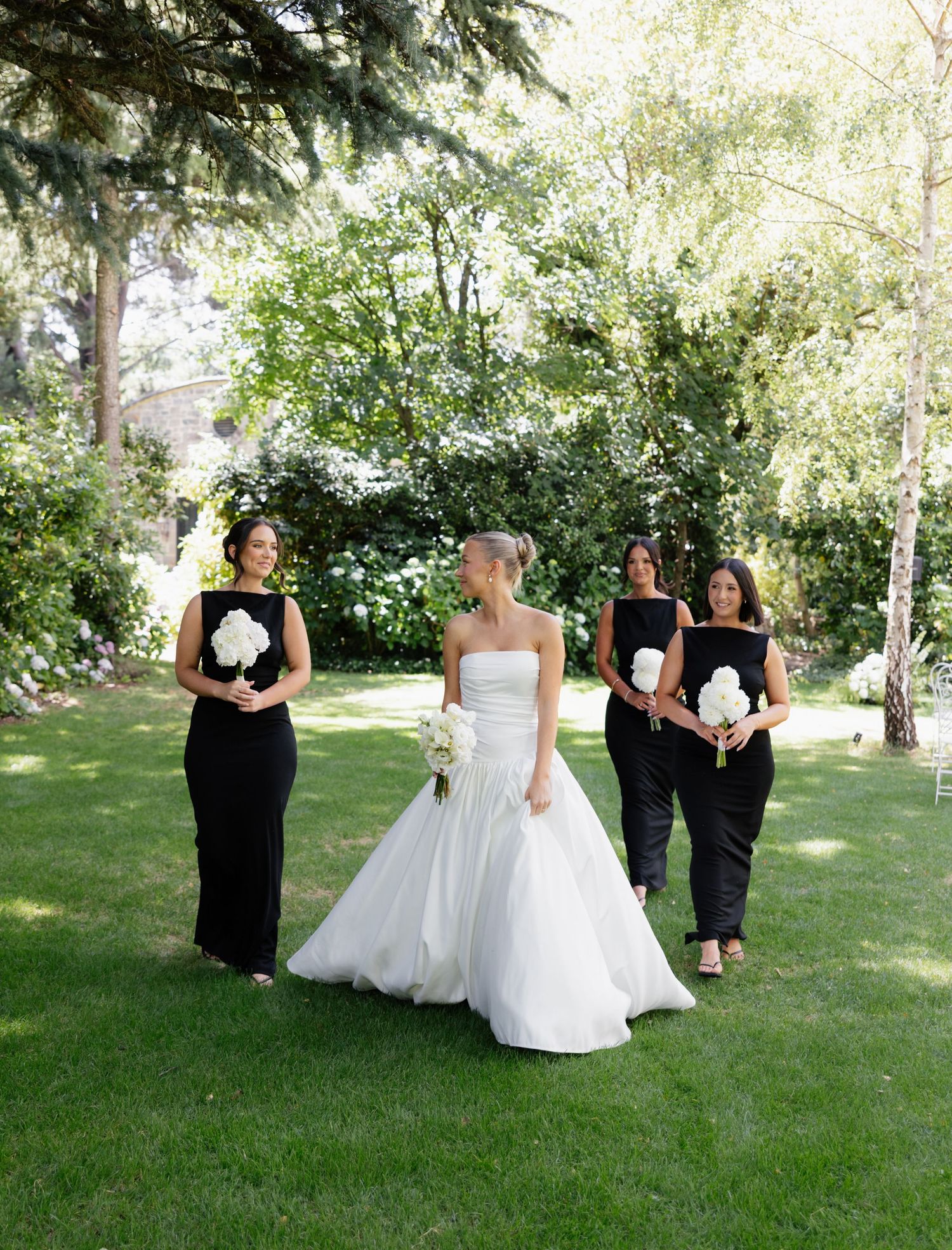 Bridal party members in black dresses gather on green grass for casual outdoor photos.