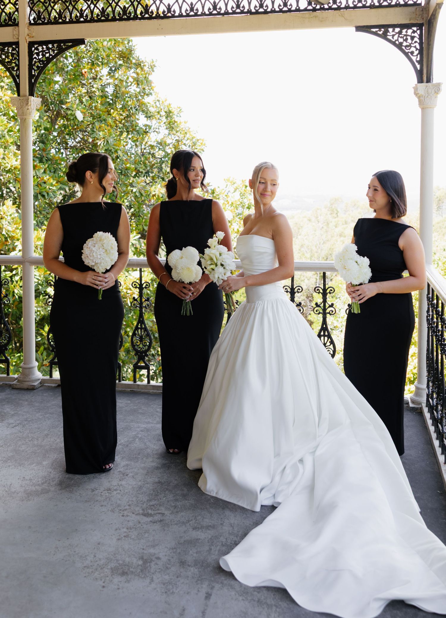 Bridesmaids in black dresses stand with bride in flowing white gown on a covered stone veranda.