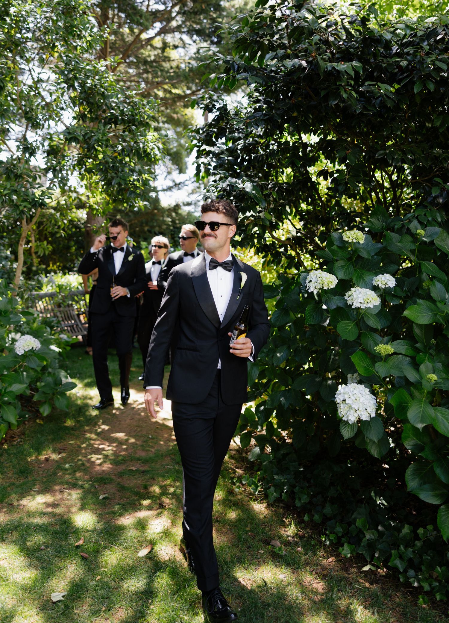 Groomsmen in black tuxedos walking along a garden path lined with white flowers and lush greenery.