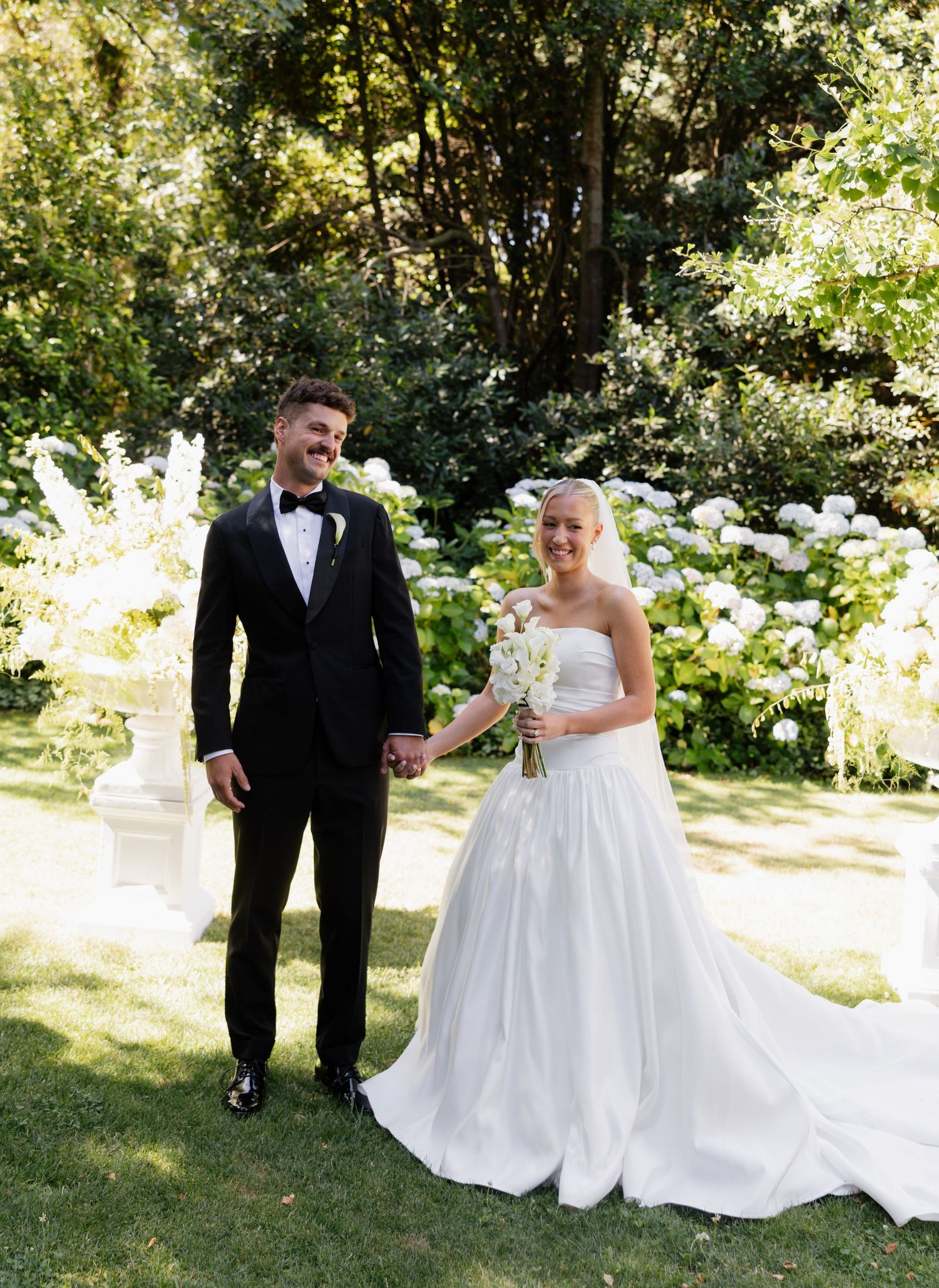 A couple in formal wedding attire shares playful moments together in a sunlit garden setting.