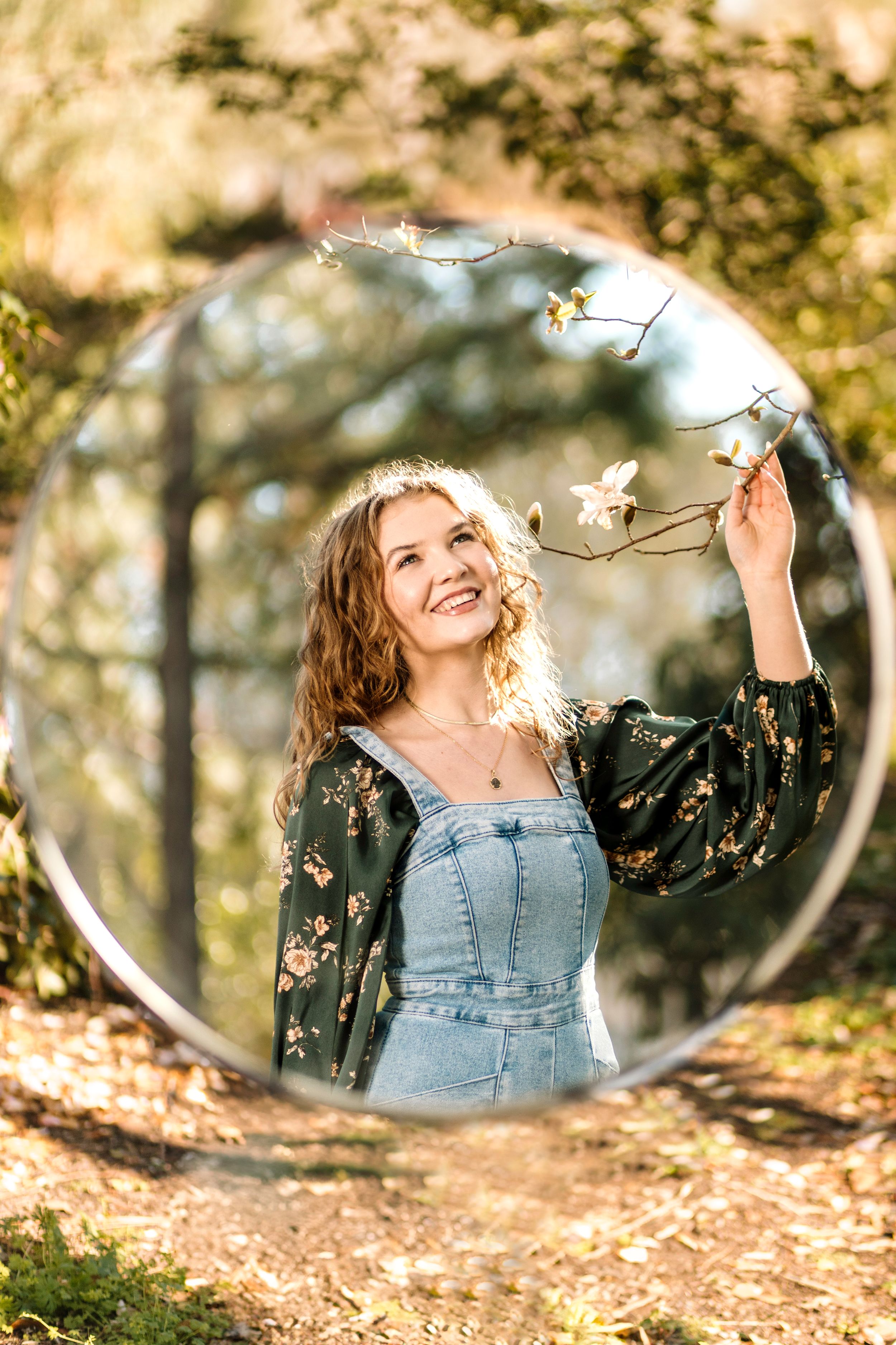 Creative portrait through circular mirror reflection shows someone in denim overalls and floral top in sunlit autumn woods.