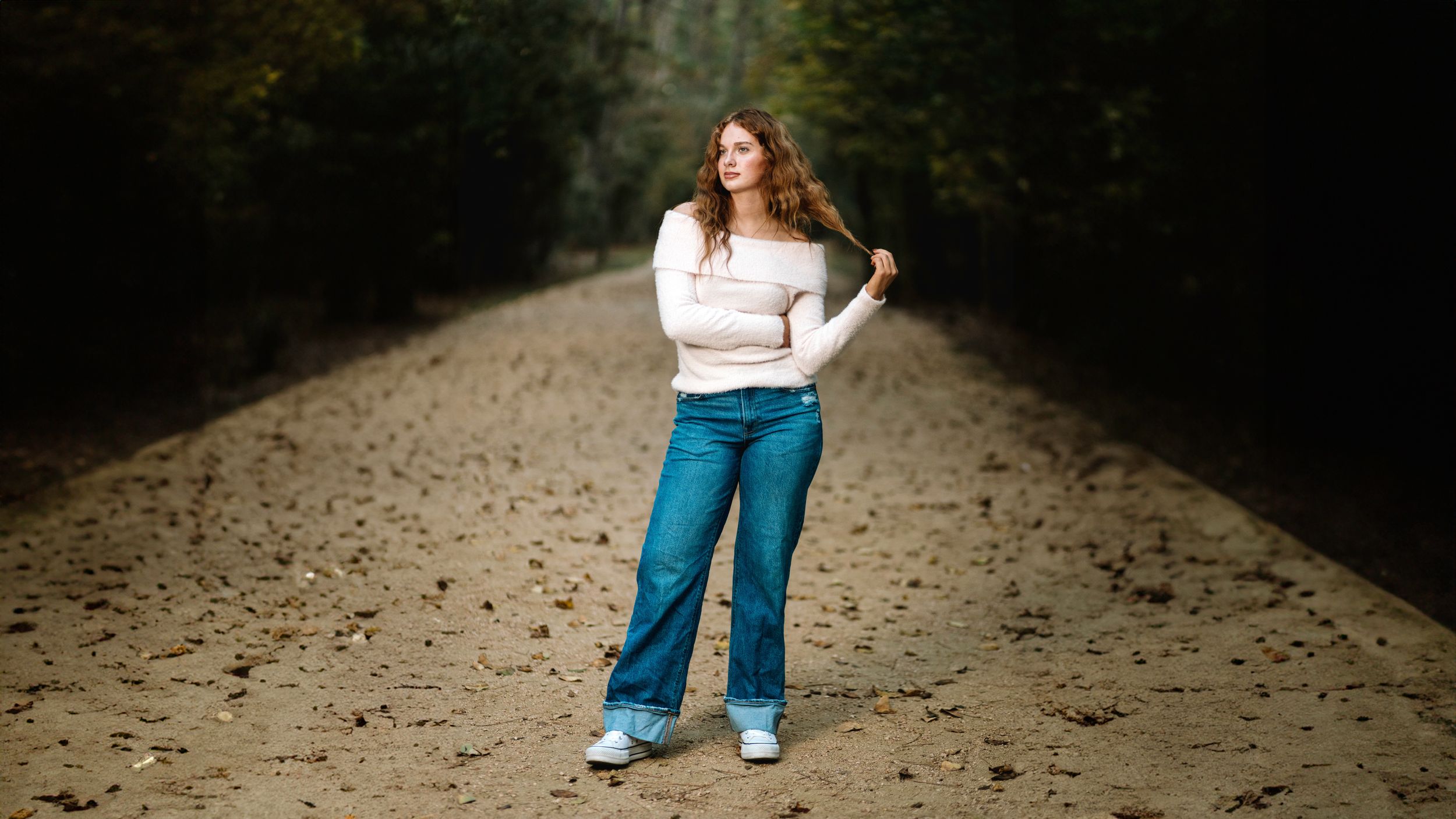 A person wearing blue jeans walks along a sandy path through a wooded area on an autumn day.