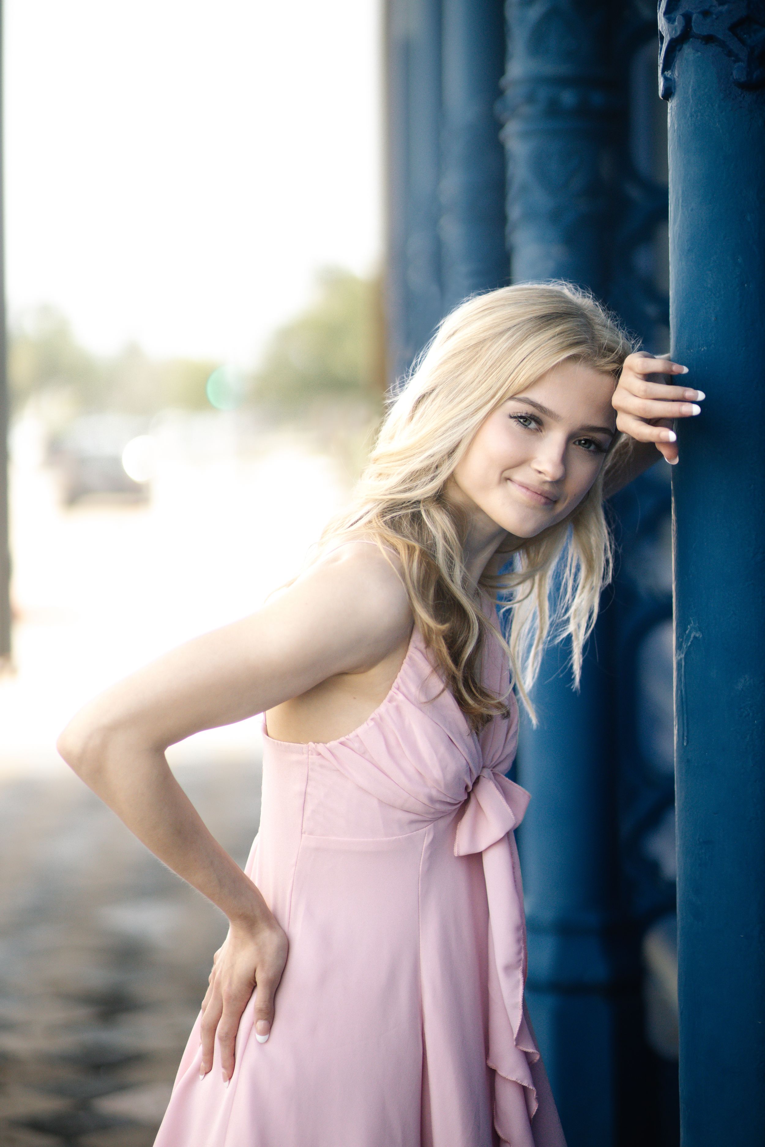 A person in a pink sleeveless dress poses gracefully against blue wooden pillars in an outdoor setting.