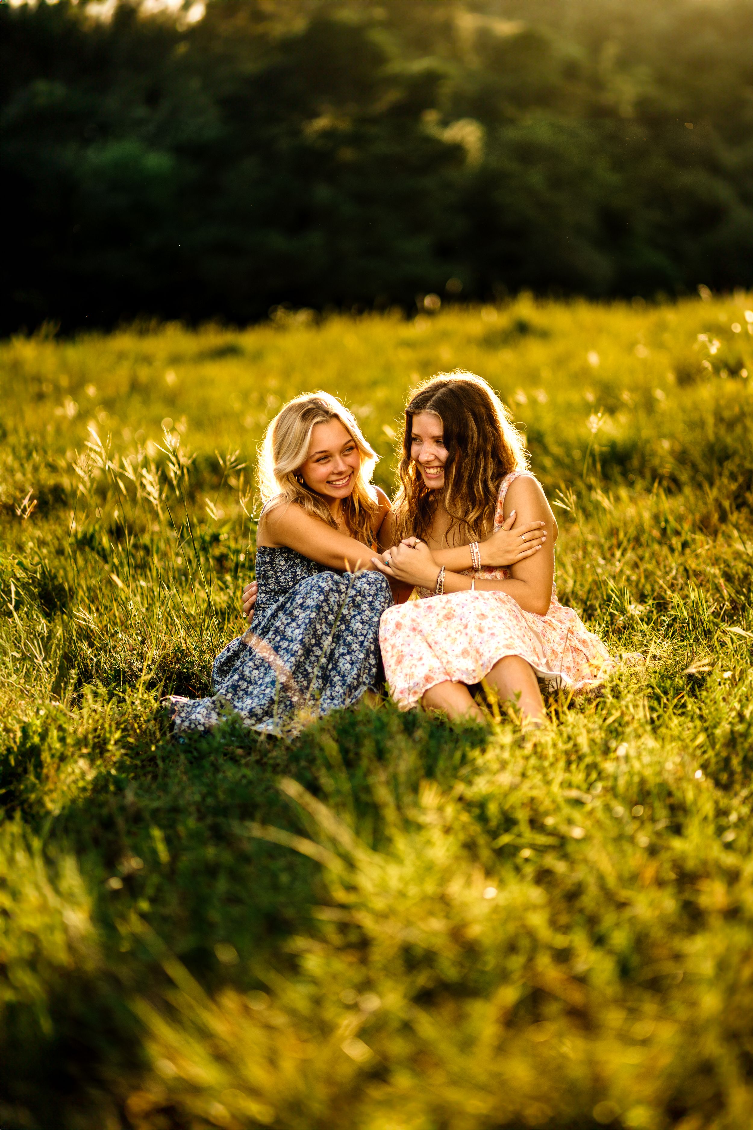 Two friends in summer dresses share a joyful moment while sitting together in a sunlit meadow at golden hour.