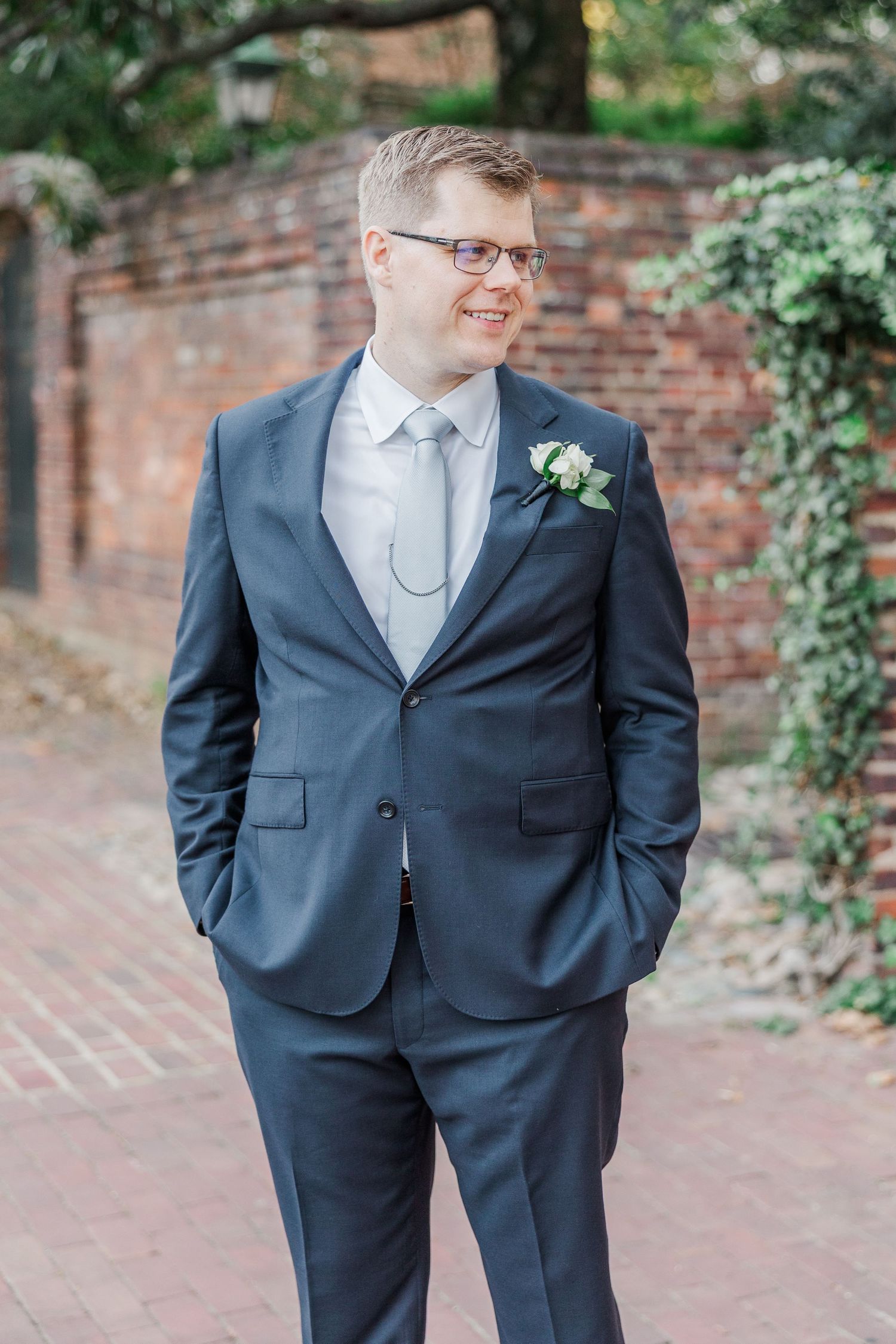 A person in a navy blue suit with a white tie stands against a brick wall with greenery in the background.