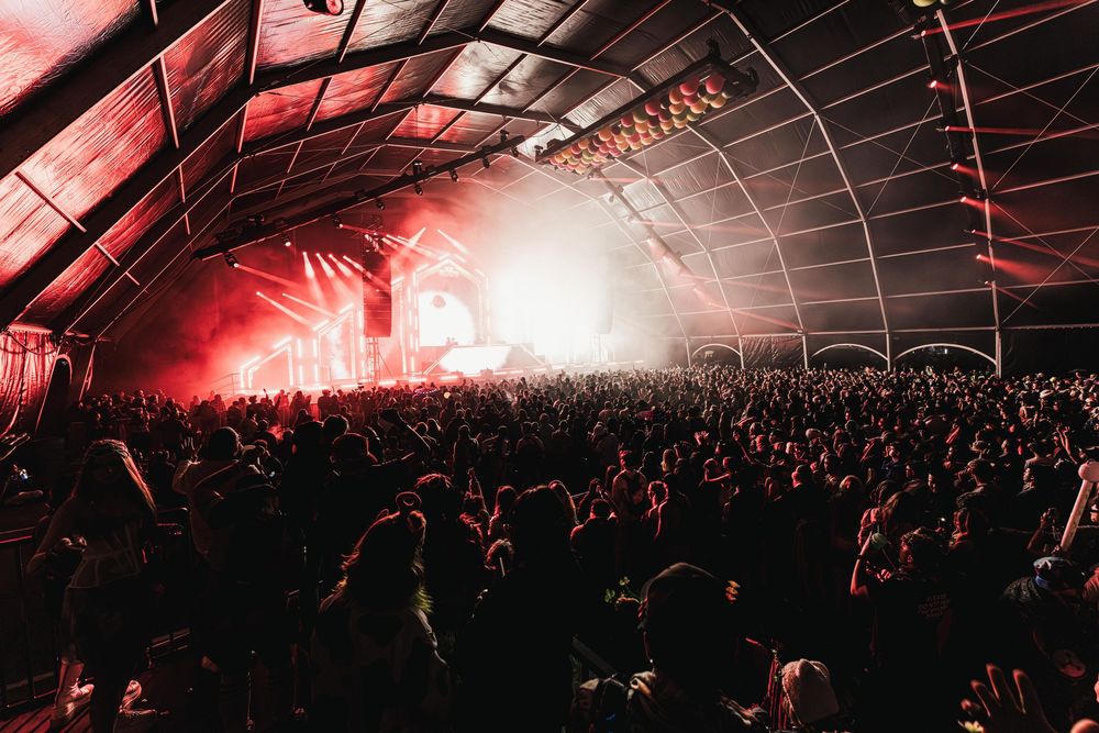 A large crowd at a music festival under a tent with dramatic red lighting and smoke effects during a nighttime performance.