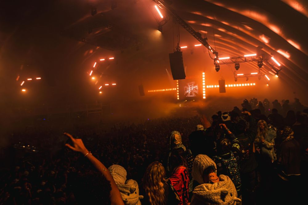 Concert crowd illuminated by orange stage lights during an energetic live music performance at a packed venue.