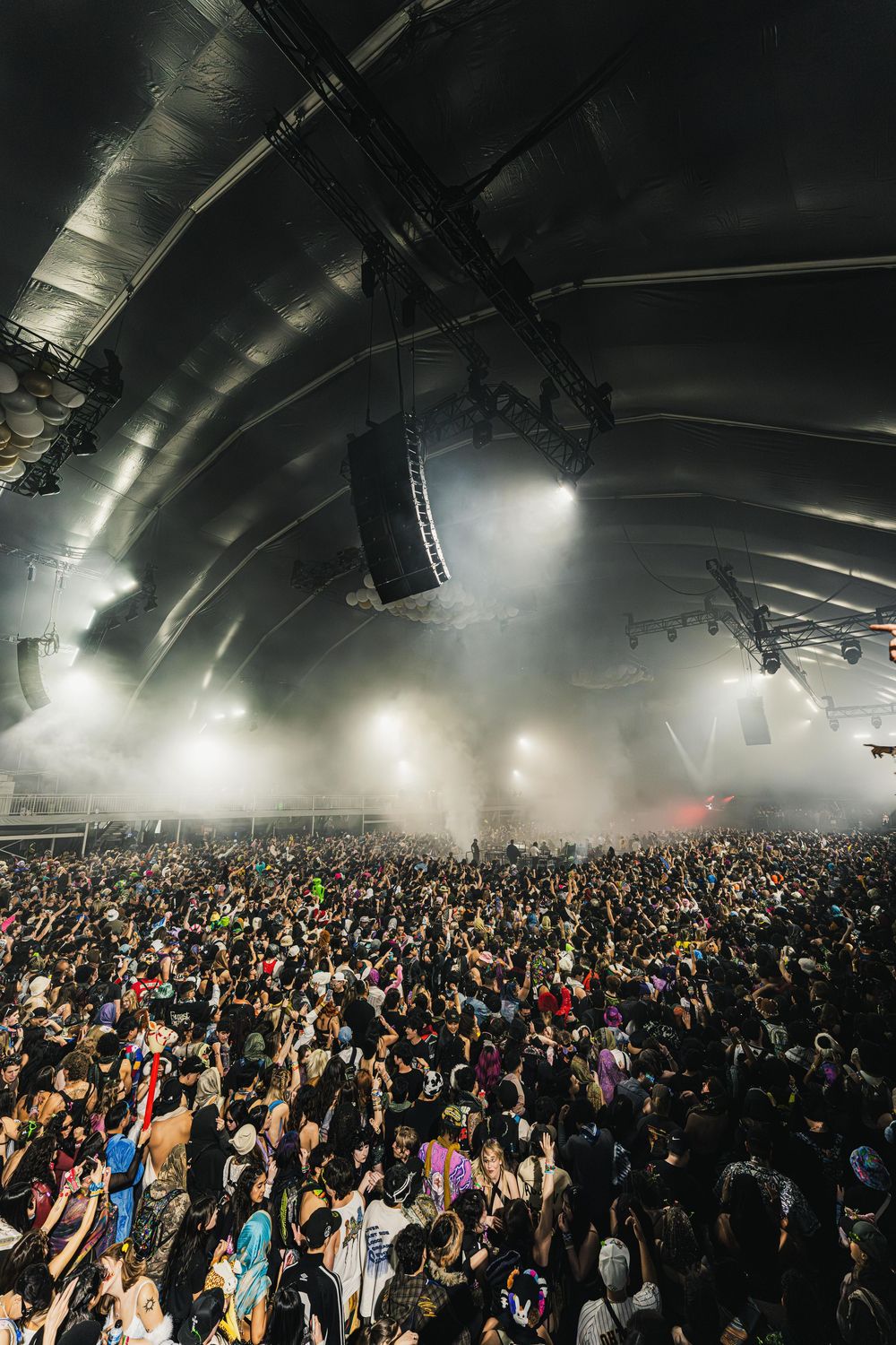 Large crowd of concert attendees fills a dark indoor venue with dramatic overhead lighting creating an atmospheric scene.