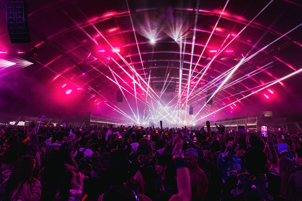 Pink and purple laser lights create geometric patterns above an energetic festival crowd at night.