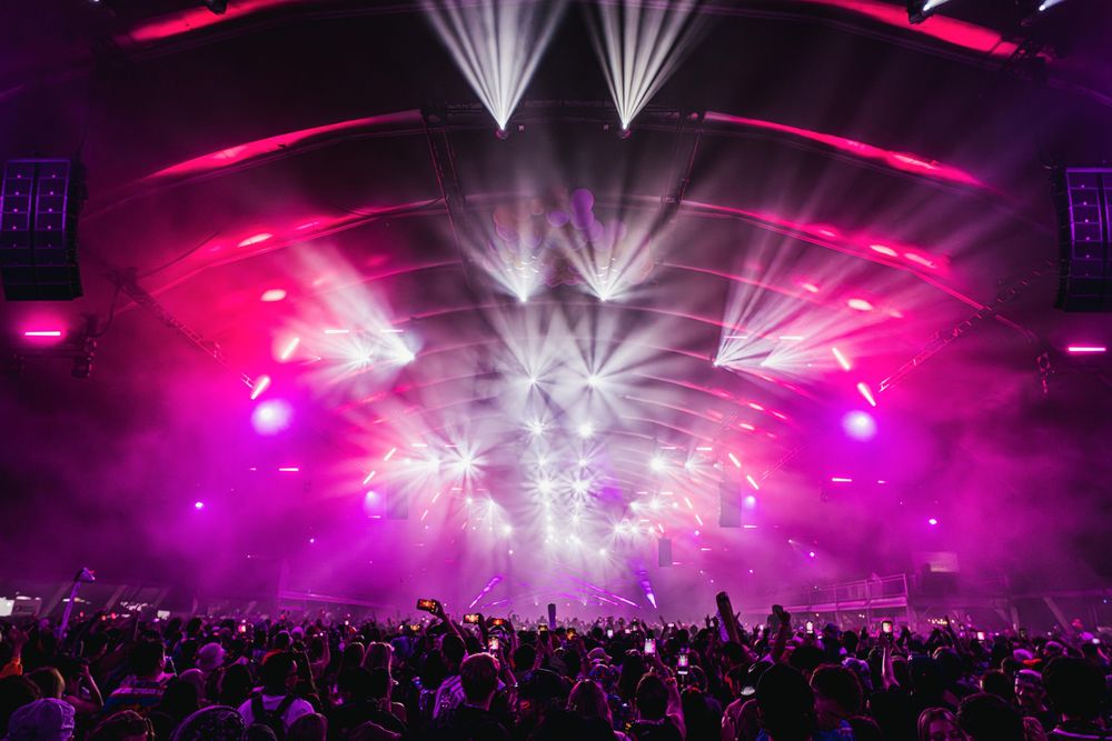 Concert venue illuminated with vibrant pink and white spotlights shining over a large crowd during a live performance.
