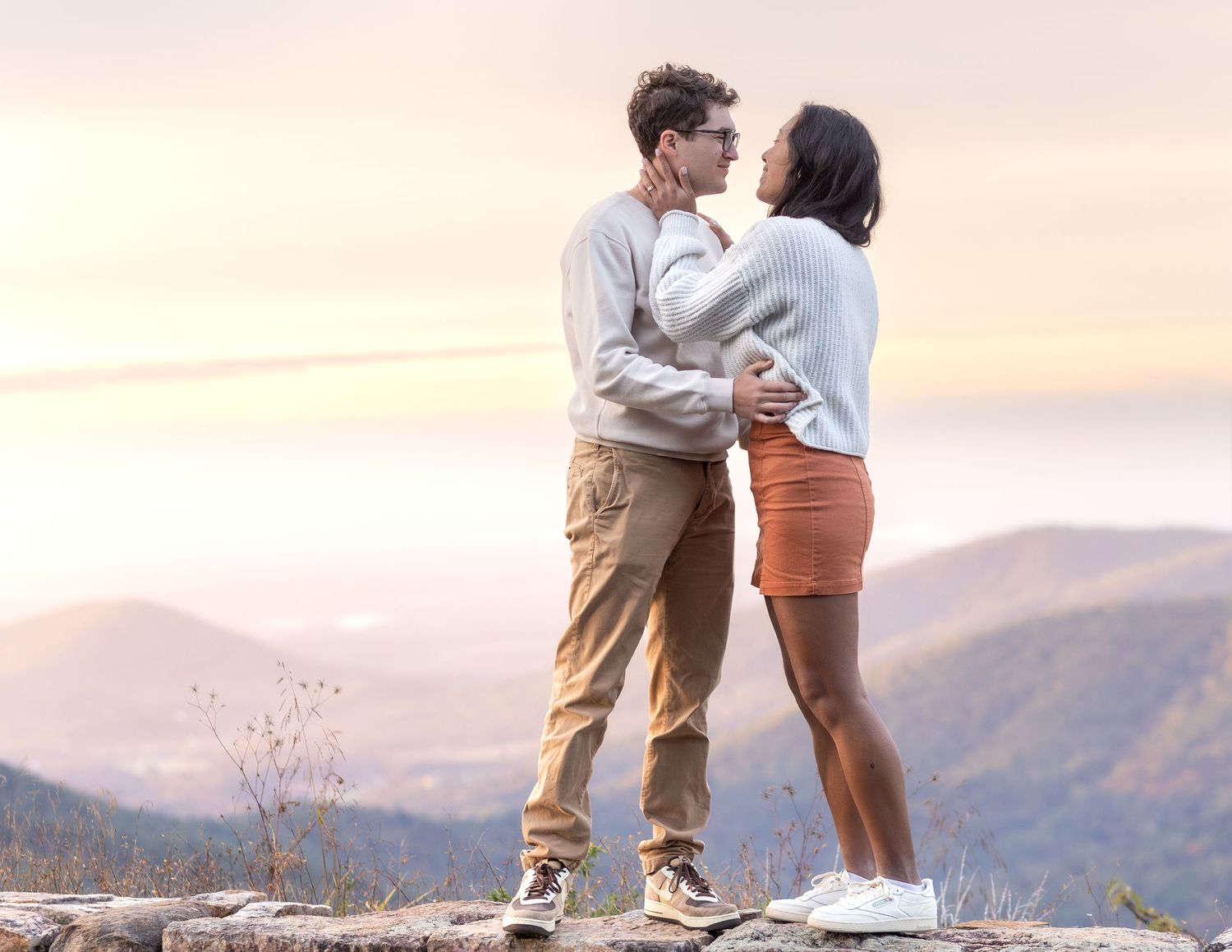 Couple embraces on mountain peak during sunset with panoramic mountain vista in background.