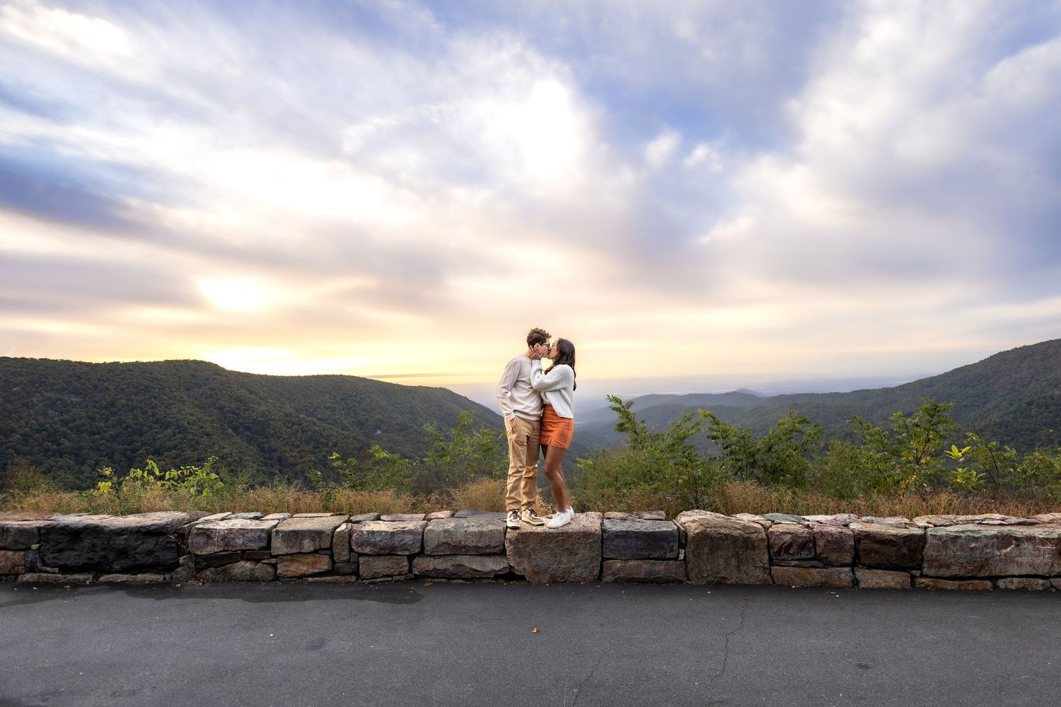 Couple embraces on wooden boardwalk overlooking scenic mountain lake at sunset with dramatic clouds in background.