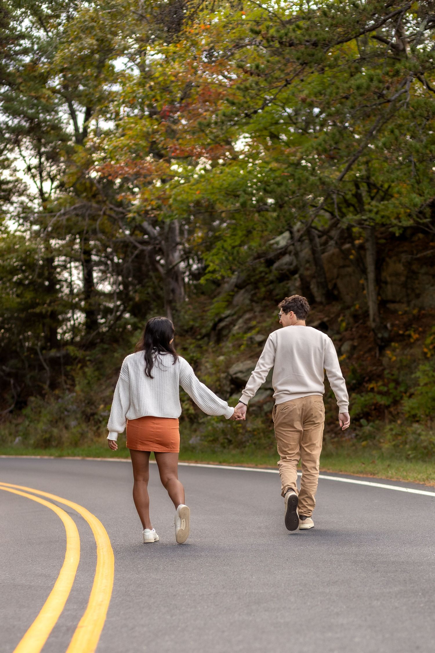 A couple walks hand in hand down a winding road surrounded by autumn trees with yellow road lines visible.