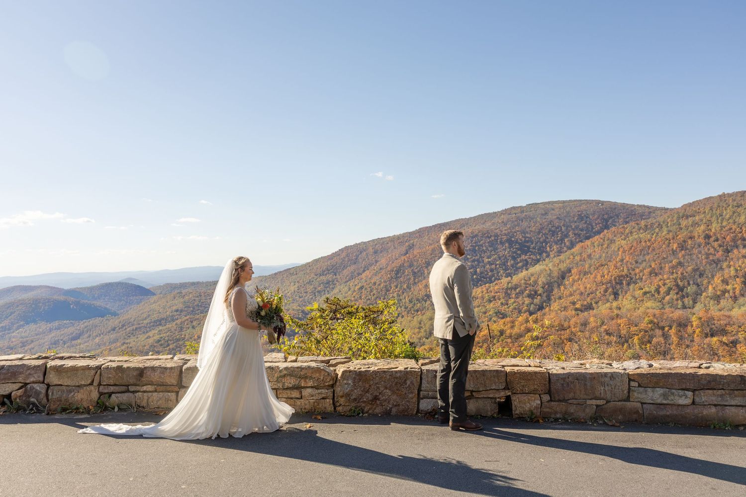 A couple in wedding attire poses together on a scenic mountain overlook with sweeping valley views at sunset.
