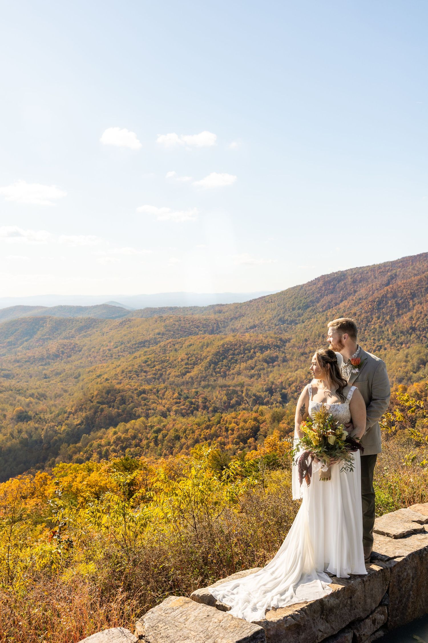 A couple shares a romantic moment at a scenic mountain overlook surrounded by vibrant autumn foliage and rolling hills.