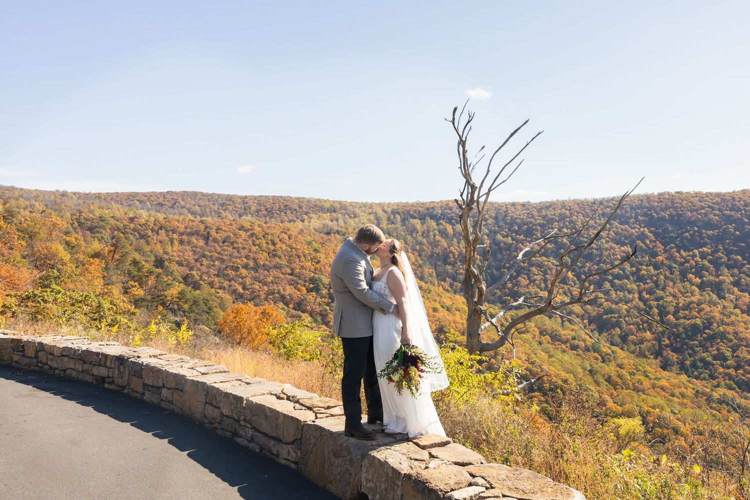 A couple stands on a scenic mountain overlook during autumn with colorful fall foliage in the background.