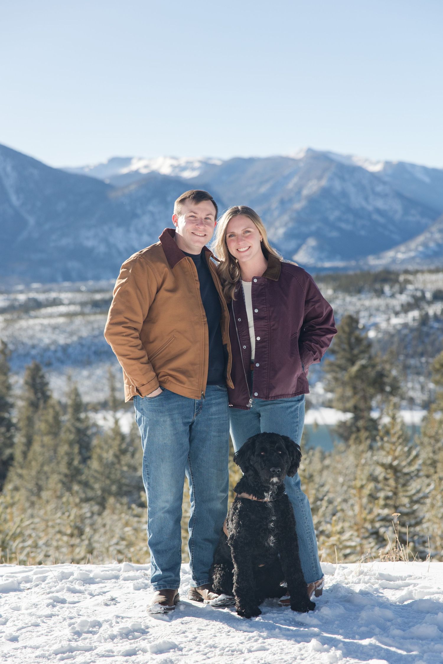 Two people walking through a snowy mountain landscape with scenic mountain peaks in the background.