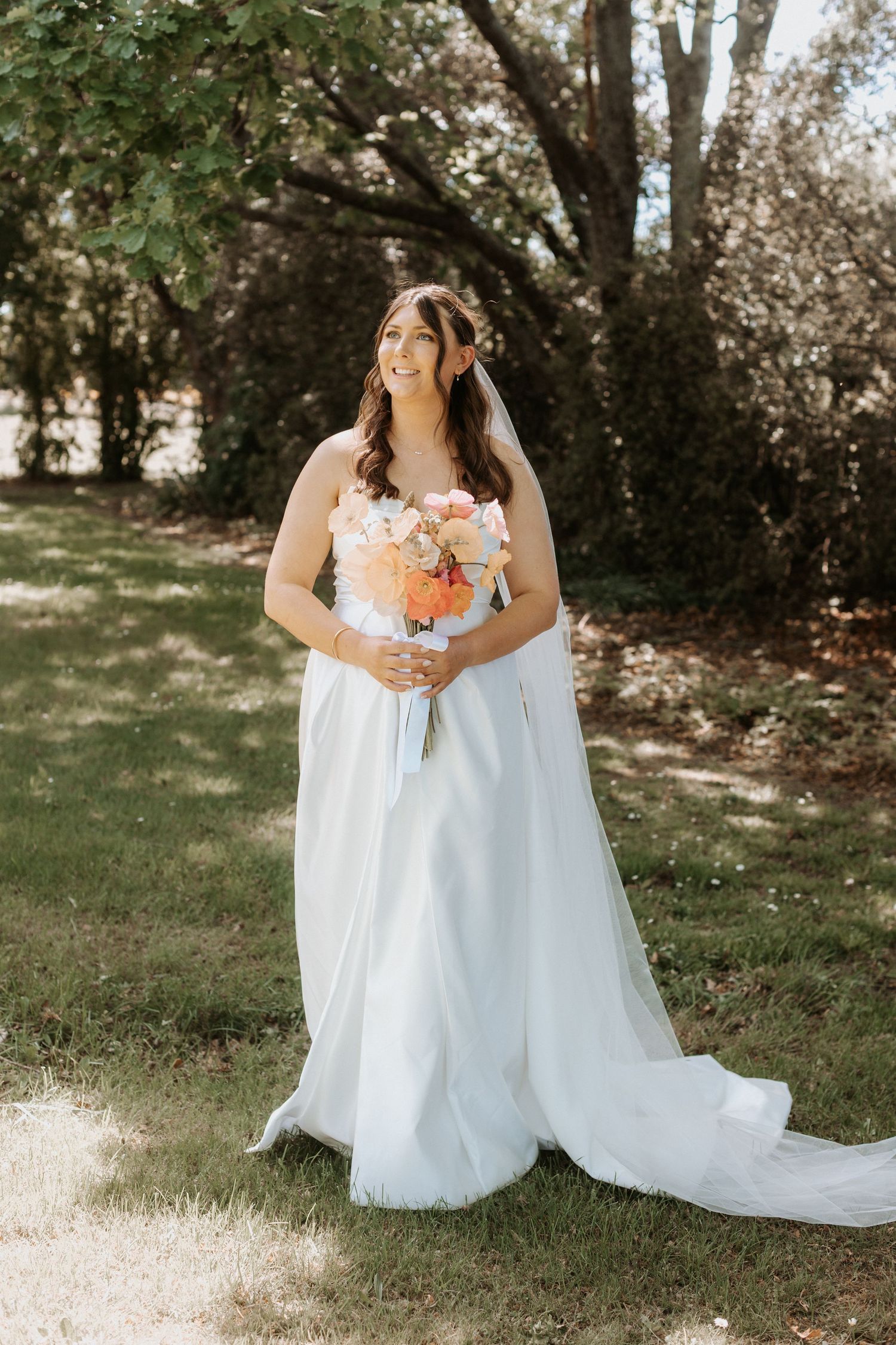 A bride in a flowing white wedding dress holds a vibrant orange and pink bouquet in a sunlit garden setting.
