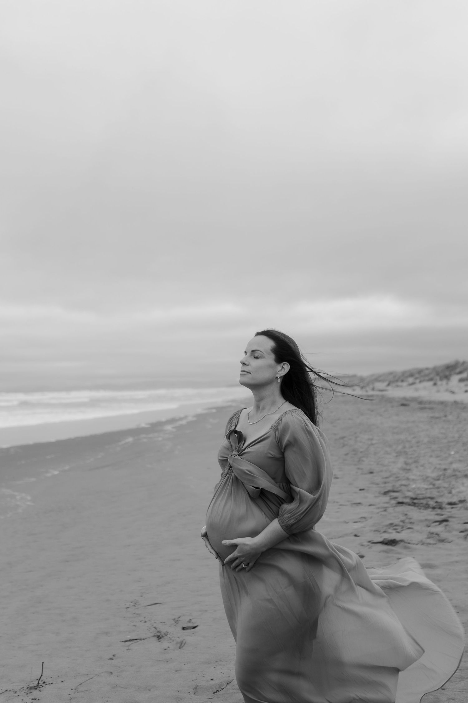 A black and white photo of a pregnant woman in a flowing dress walking along a misty beach shoreline.