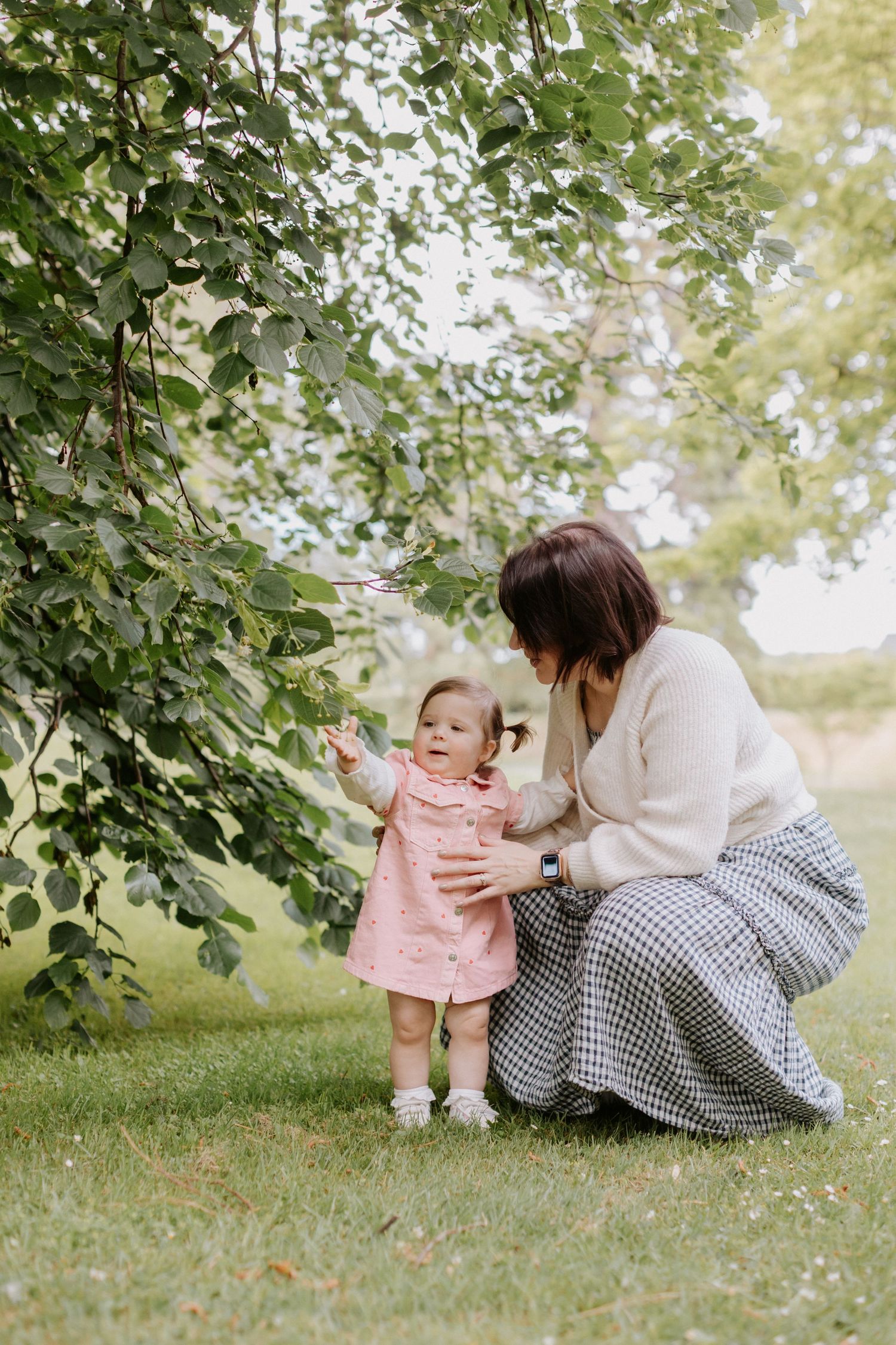 A tender outdoor moment between parent and child exploring nature together under green leafy trees on a sunny day.