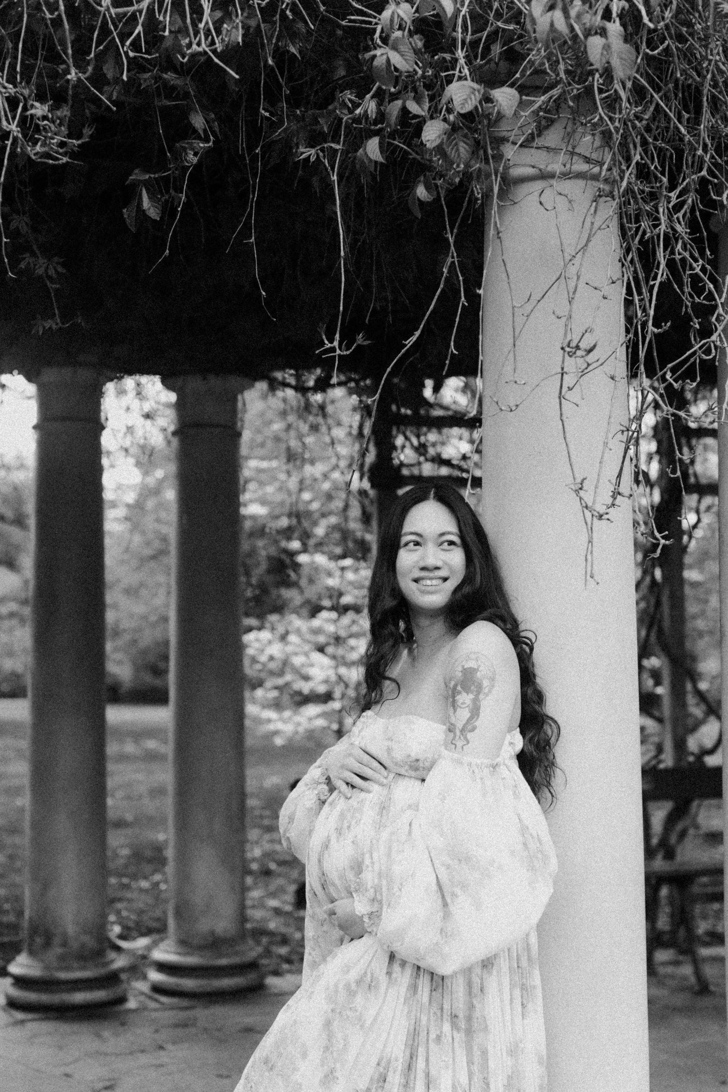 A black and white portrait of someone in a flowing white lace dress standing by classical columns with hanging vines.