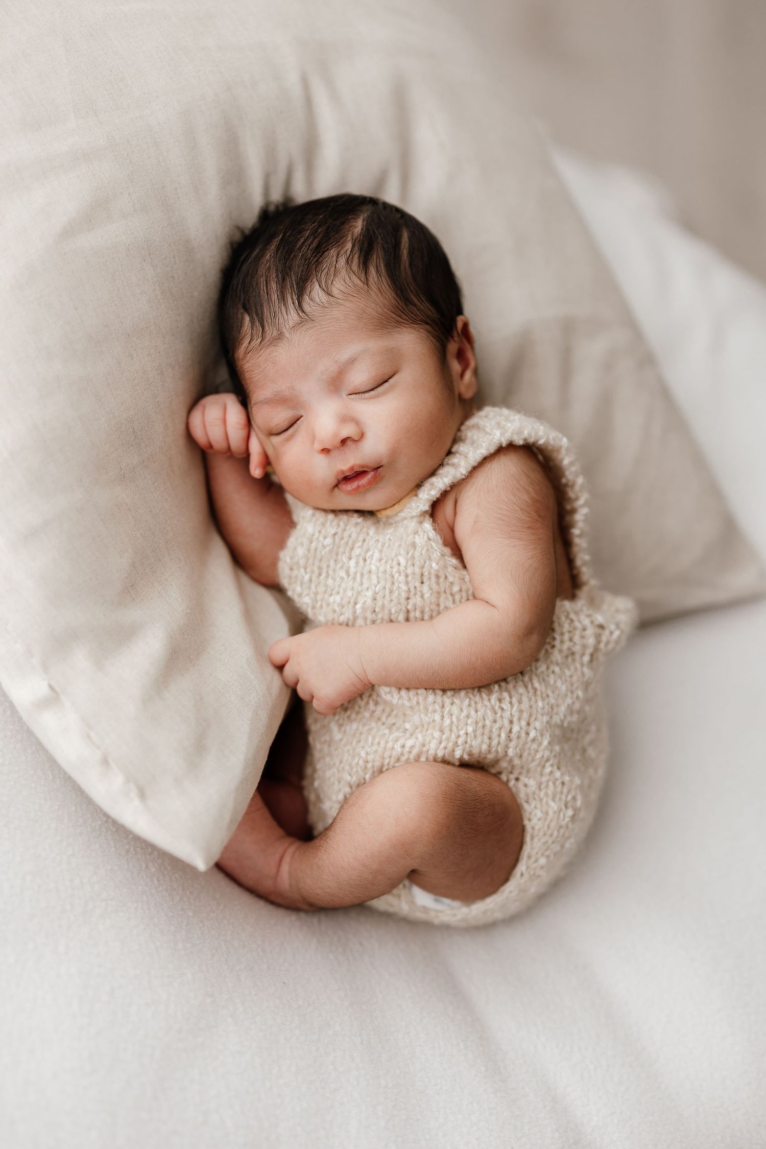 A sleeping newborn baby wrapped in a soft cream knit blanket lies peacefully on white bedding.