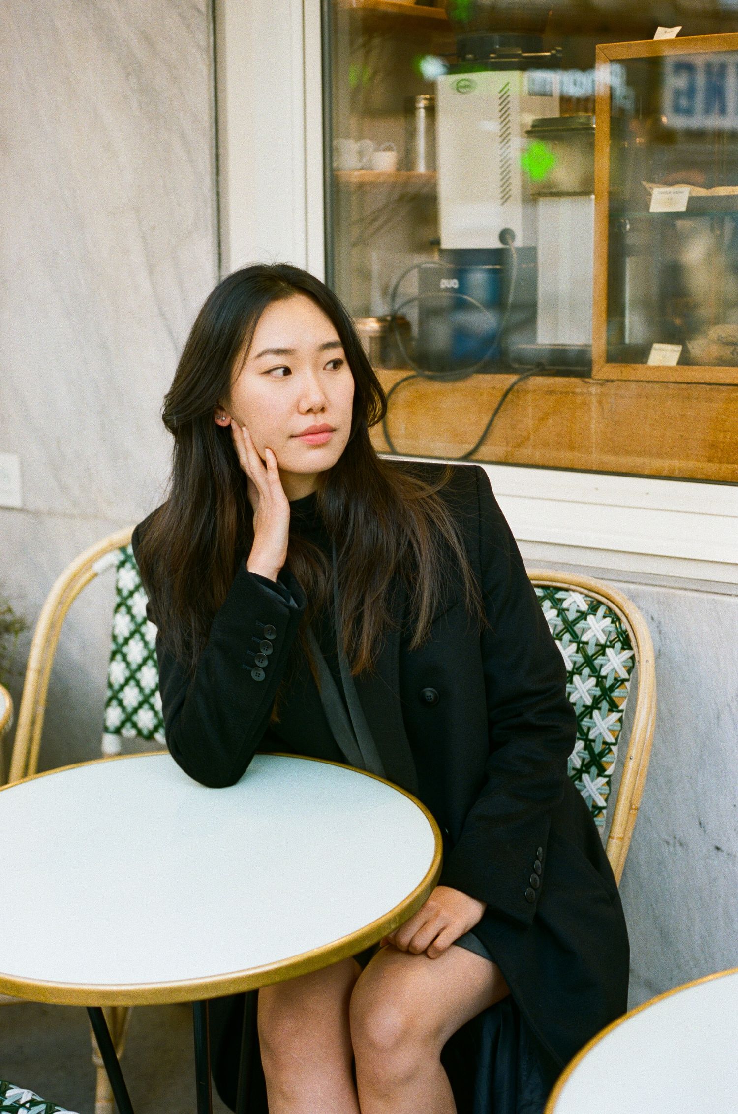 A person in a black coat sits at an outdoor cafe table with a white round top near a window.
