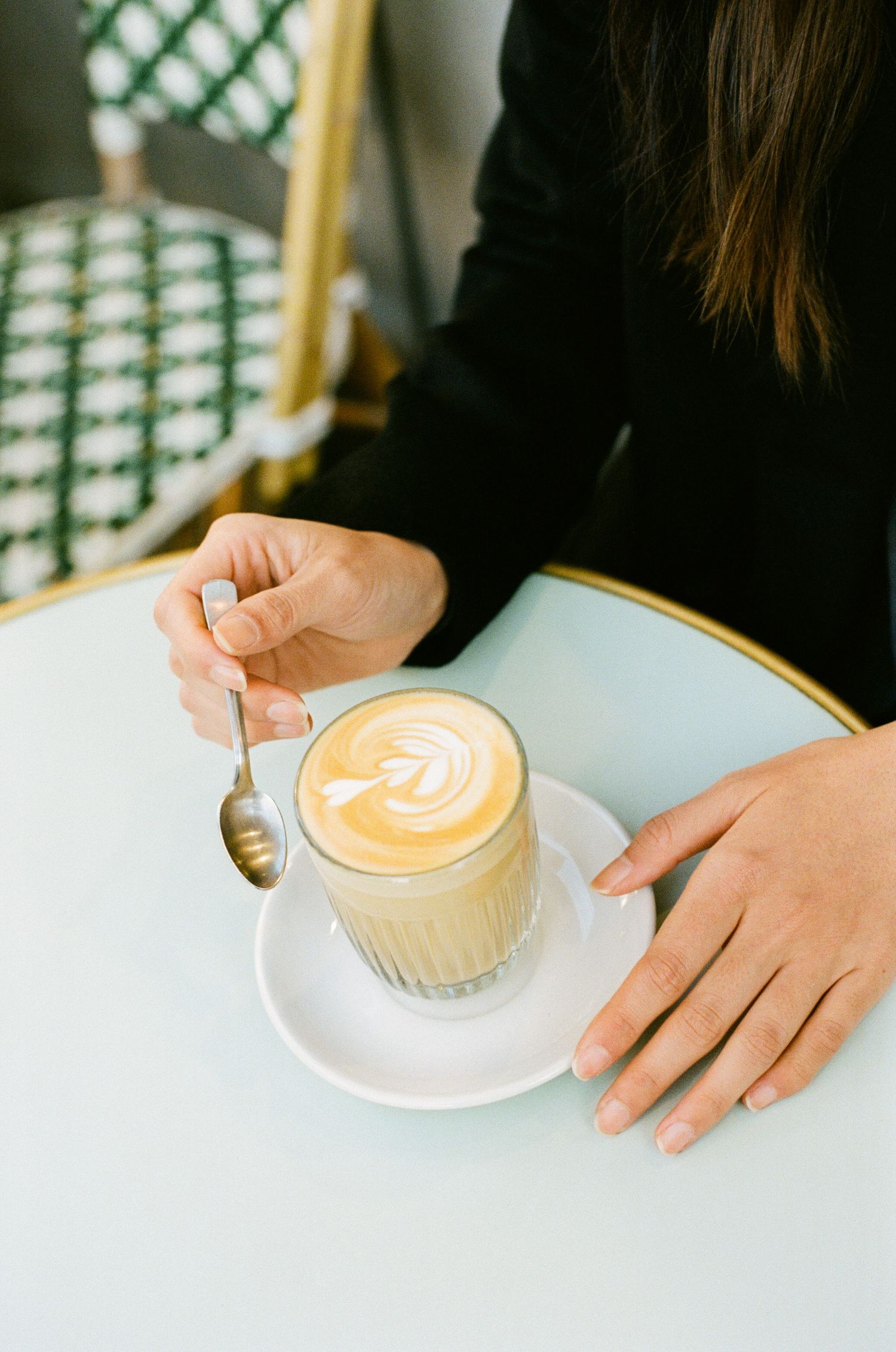 Someone stirring a latte with beautiful leaf art pattern in a glass cup on white table at cafe.